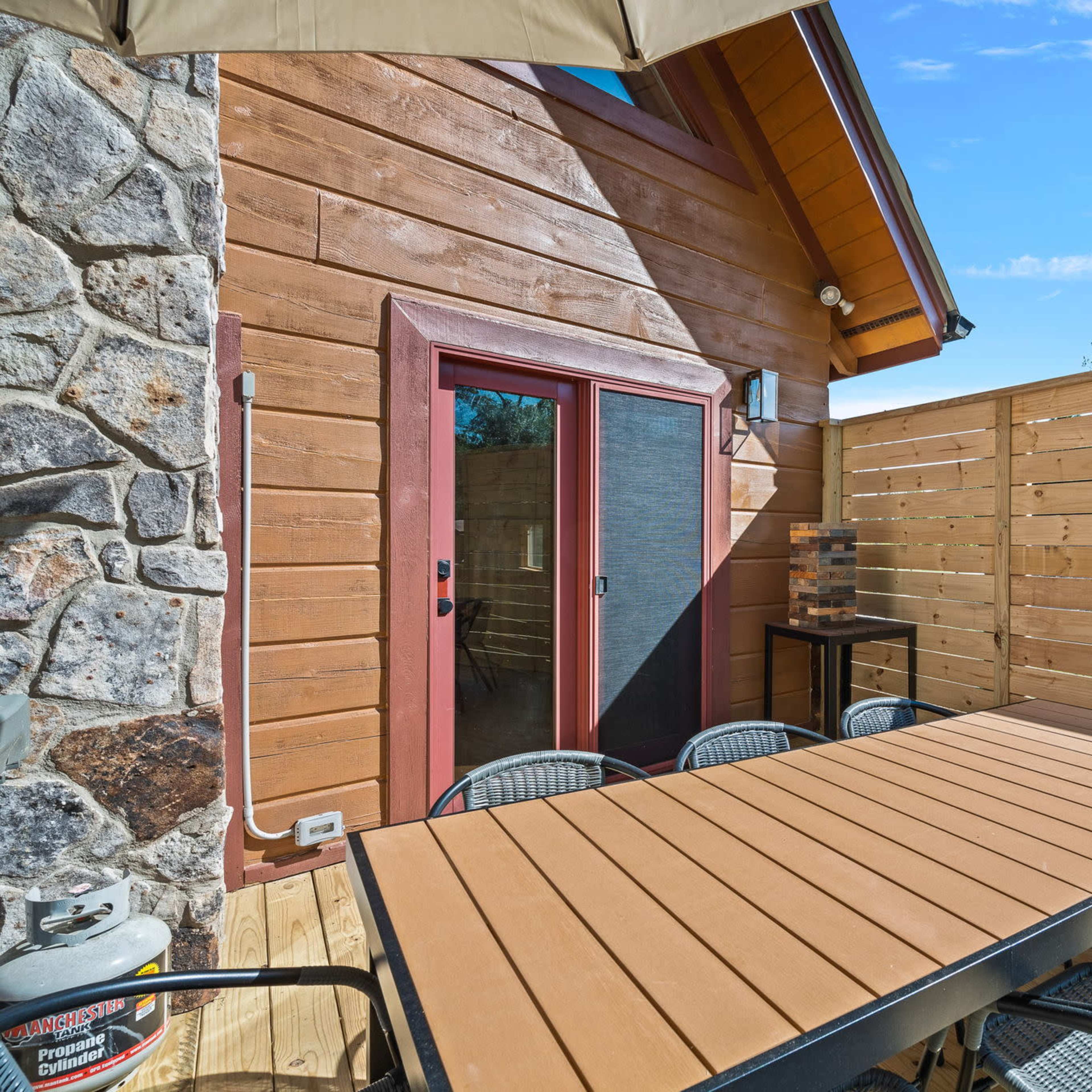 A wooden patio features a large table surrounded by chairs next to a stone wall and a fenced area with a door leading indoors.