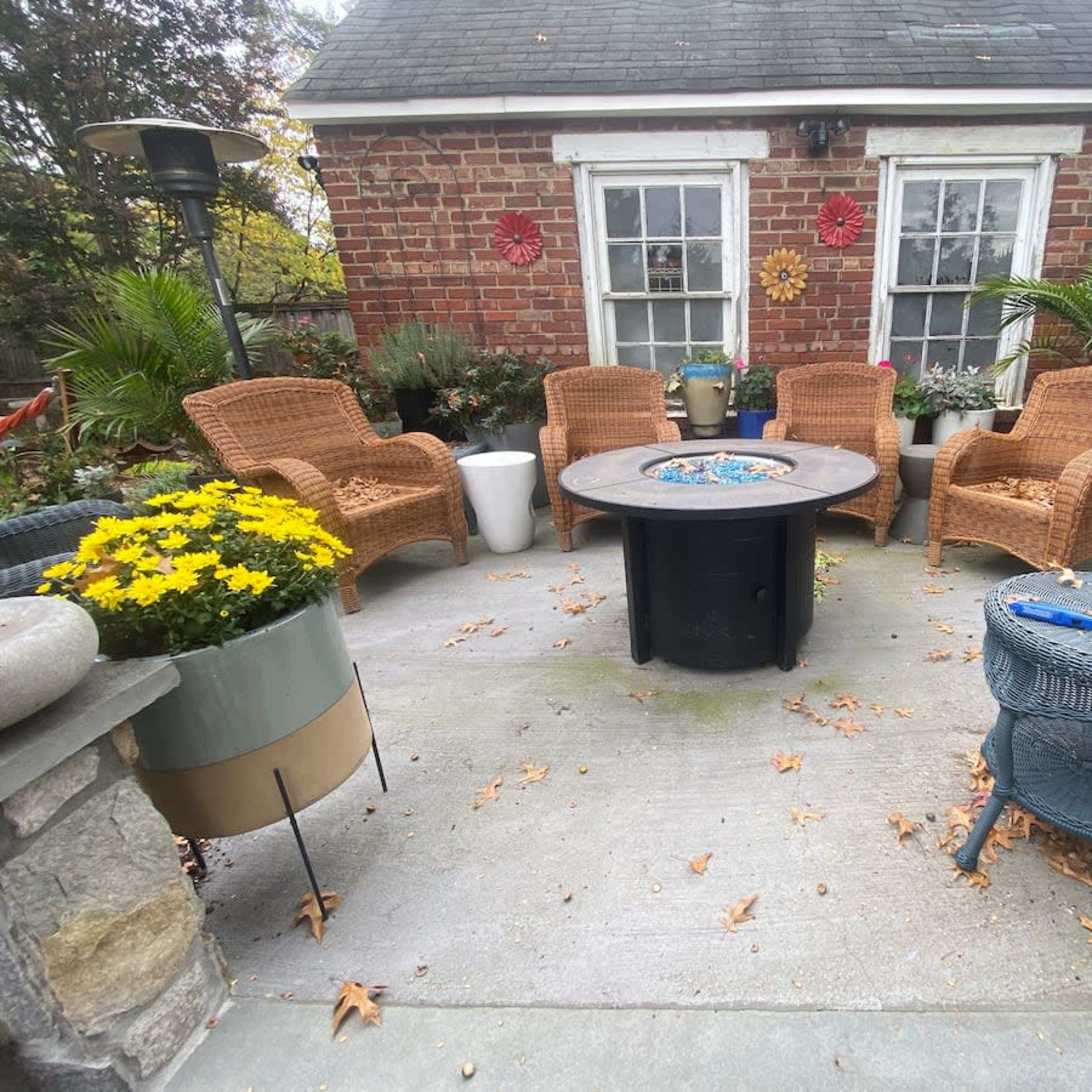 A patio with wicker chairs arranged around a circular fire pit, surrounded by potted plants and flowers.