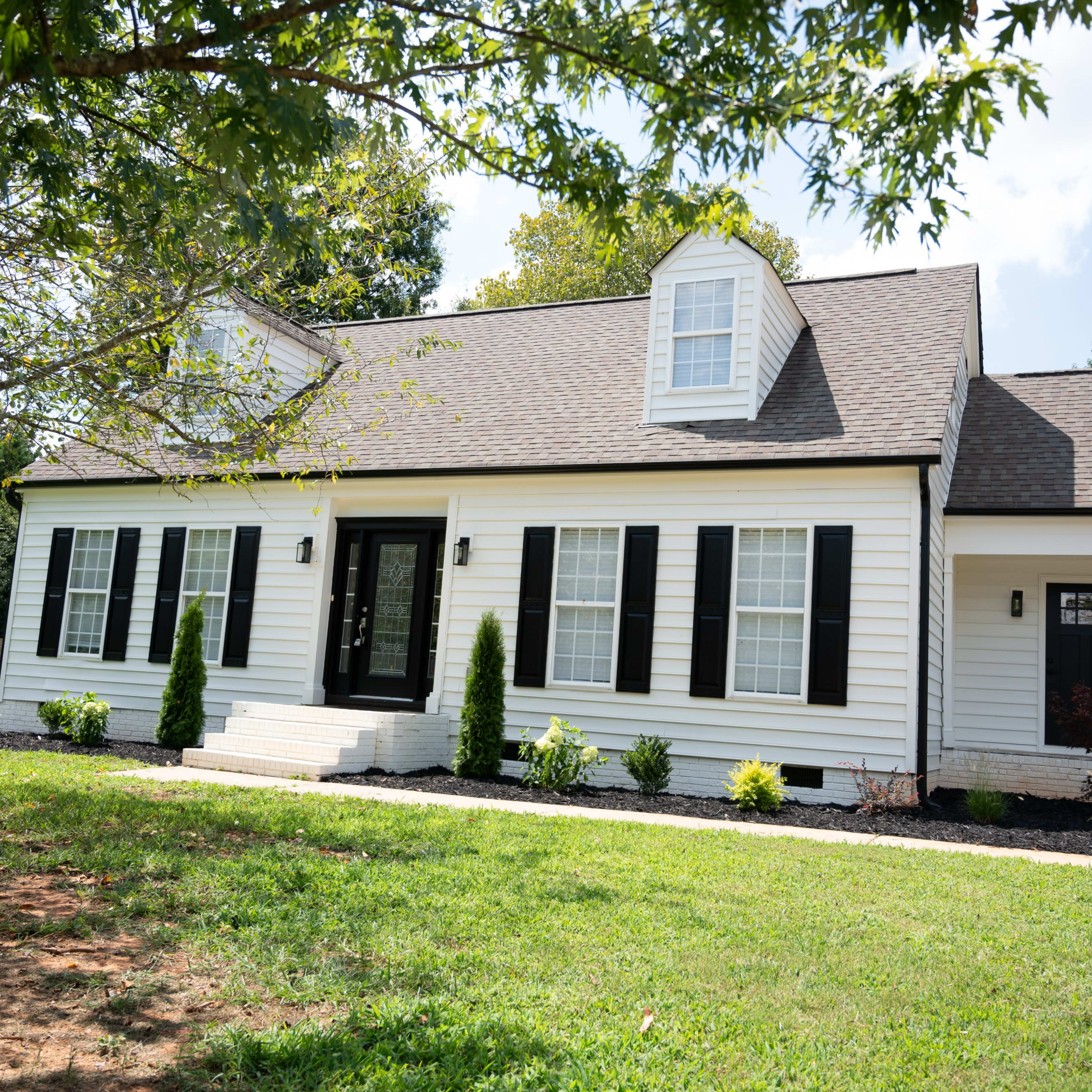 A white, single-story house with black shutters and a front porch is set on a grassy lawn surrounded by trees.