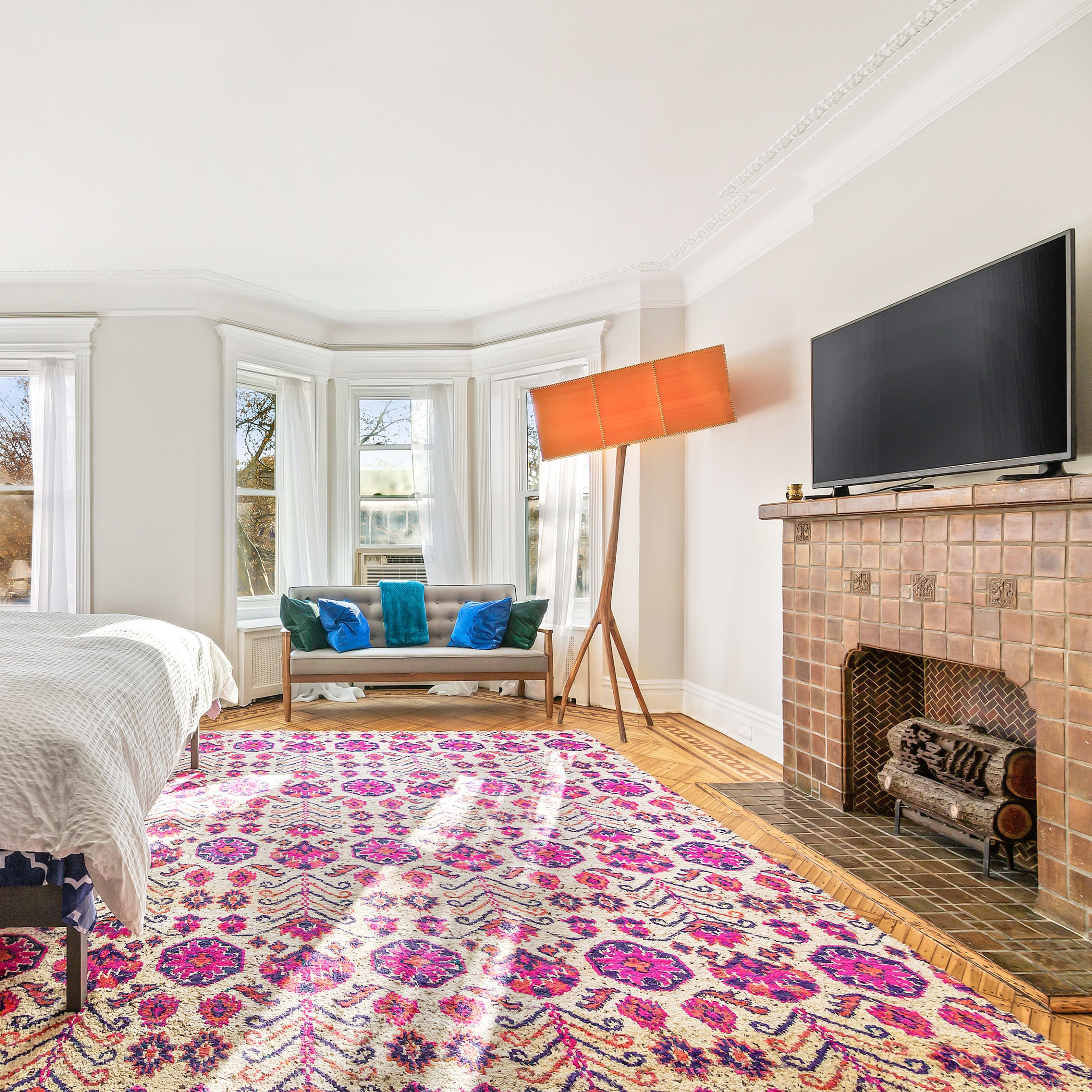 The bedroom features a bed with a patterned blanket, a decorative rug, a modern lamp, and a television mounted above a tiled fireplace, with large windows allowing natural light to fill the space.