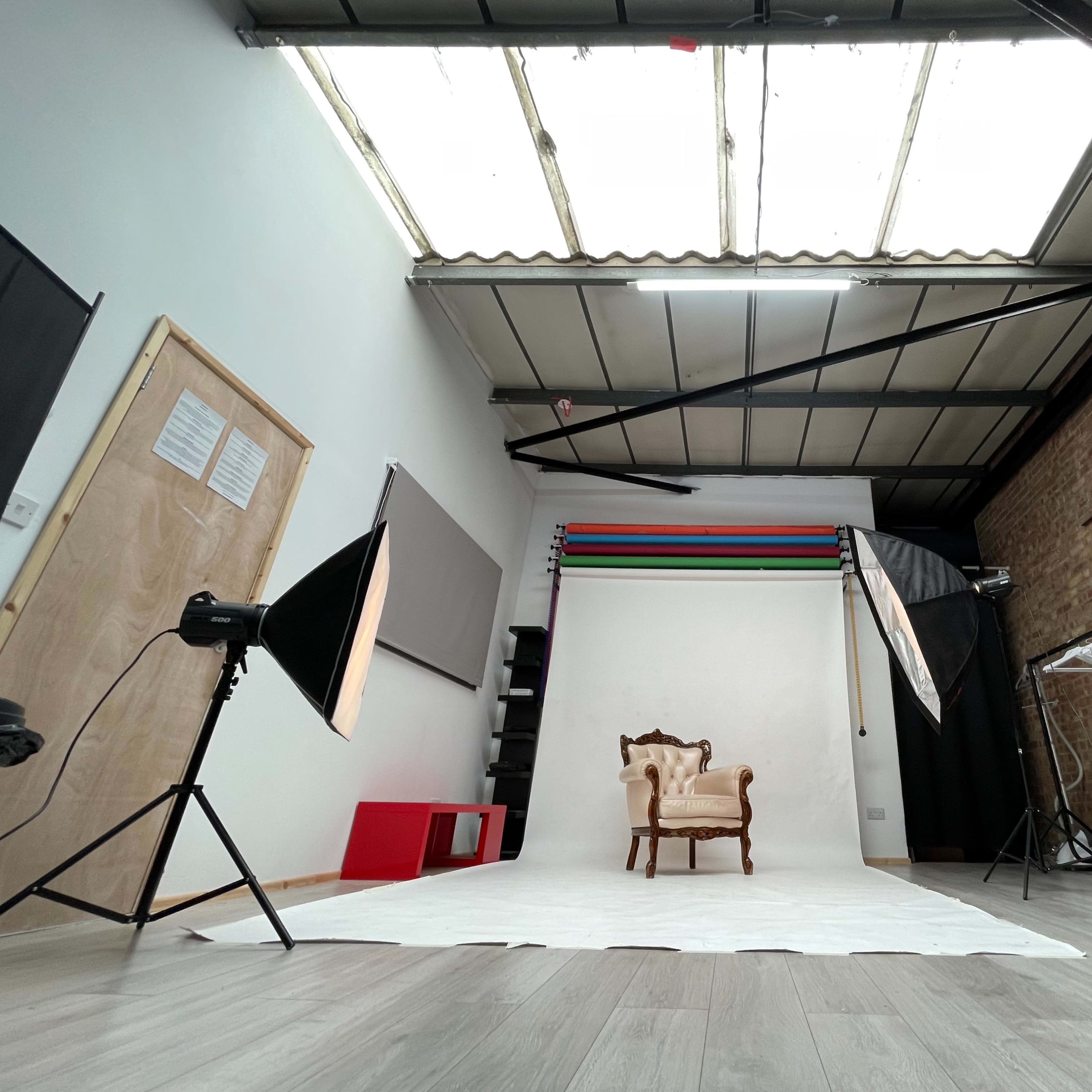 A photography studio with a vintage chair positioned on a white backdrop, surrounded by lighting equipment and a brick wall.