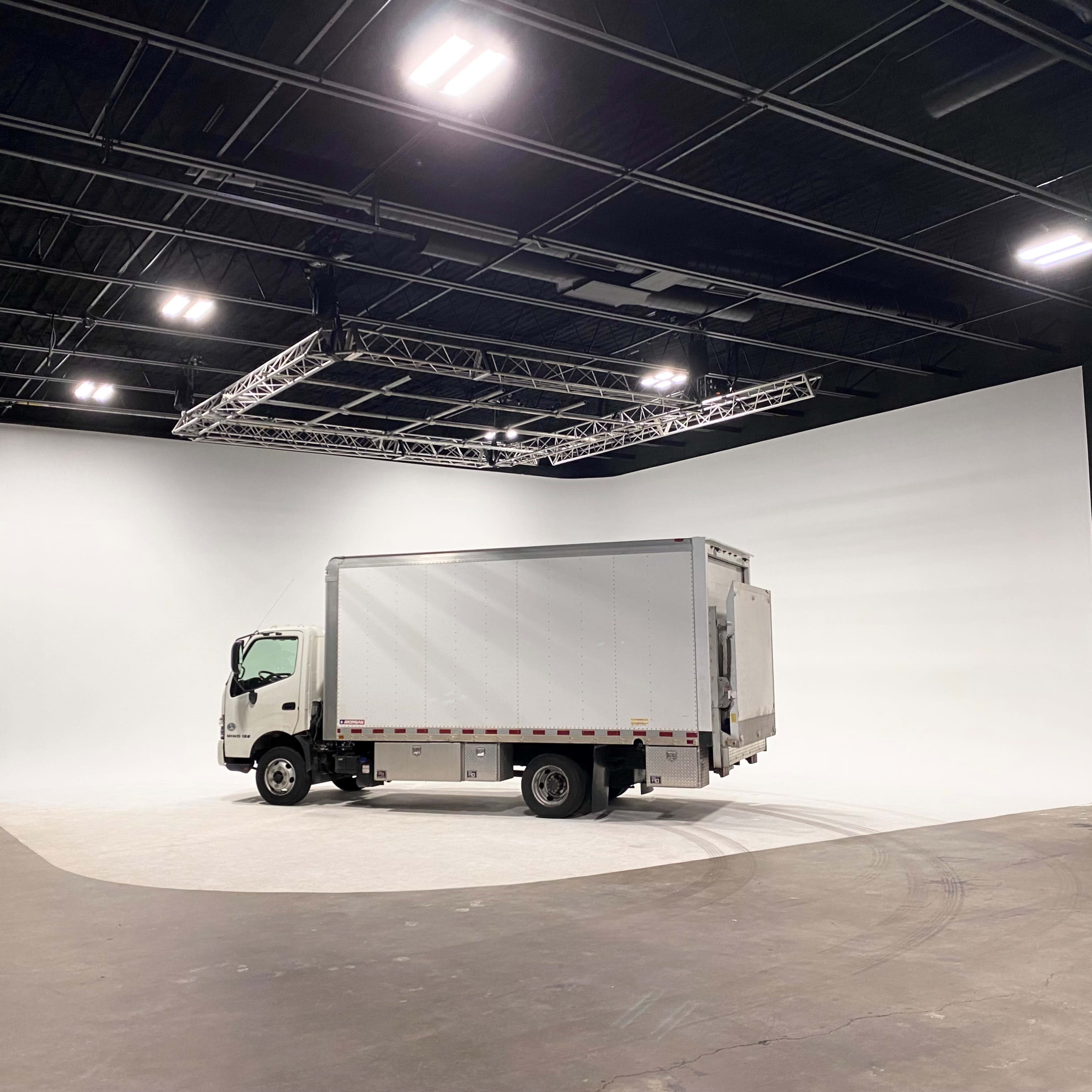A white delivery truck is parked in a spacious studio with a curved white backdrop and industrial lighting.