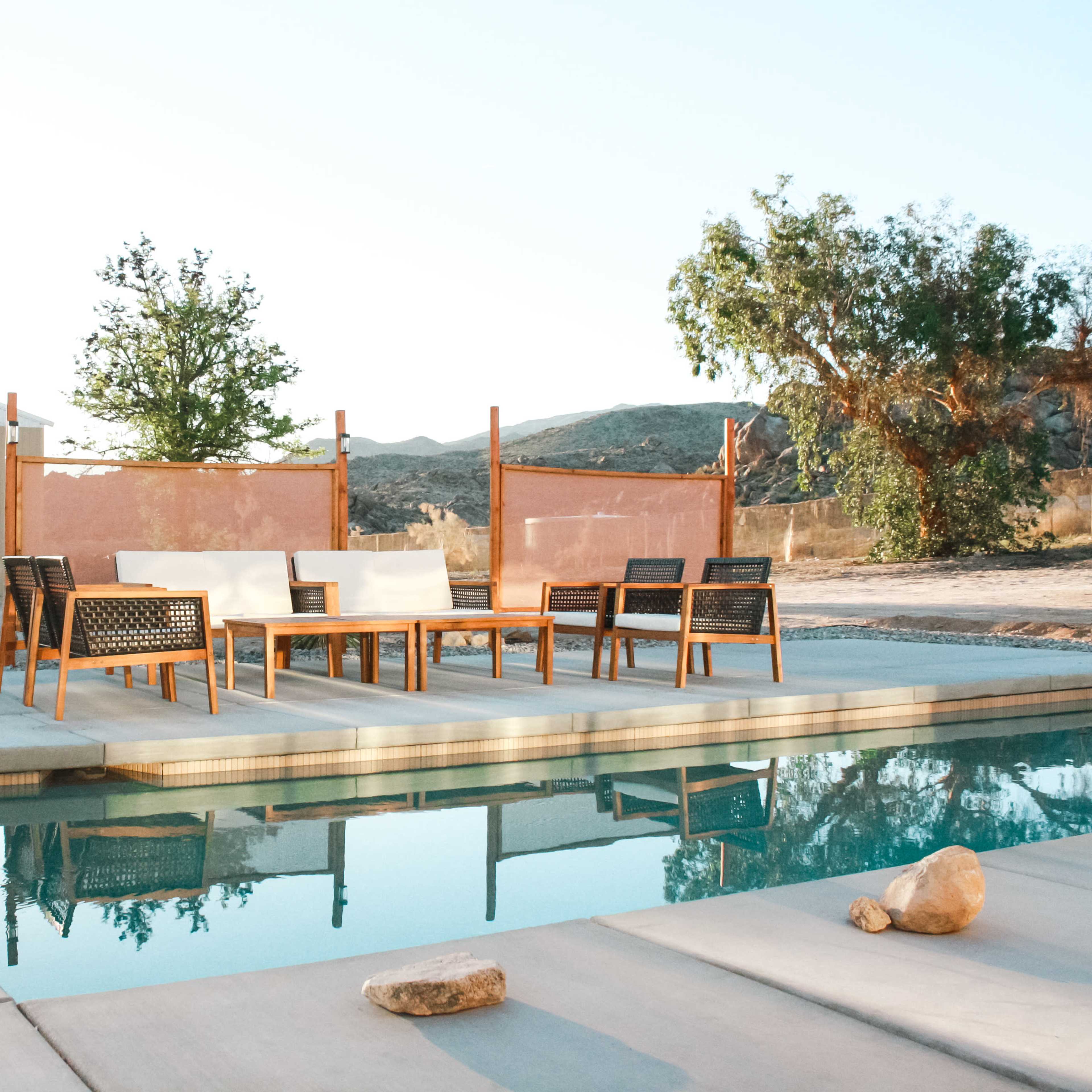 The scene features a swimming pool surrounded by wooden patio furniture, with a desert landscape and a building in the background under a clear sky.