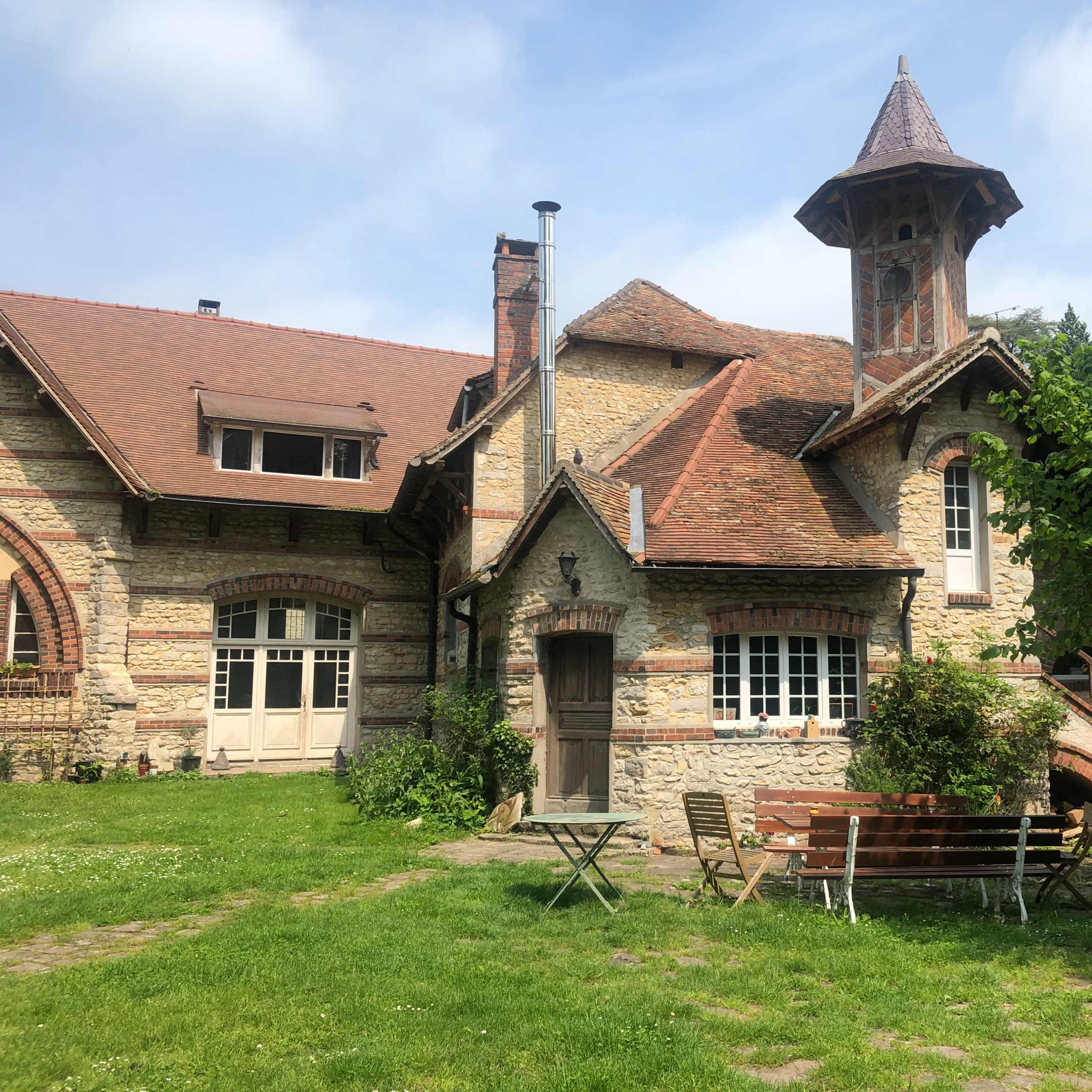 A stone house with a tower features a sloped roof and is surrounded by a garden with seating.