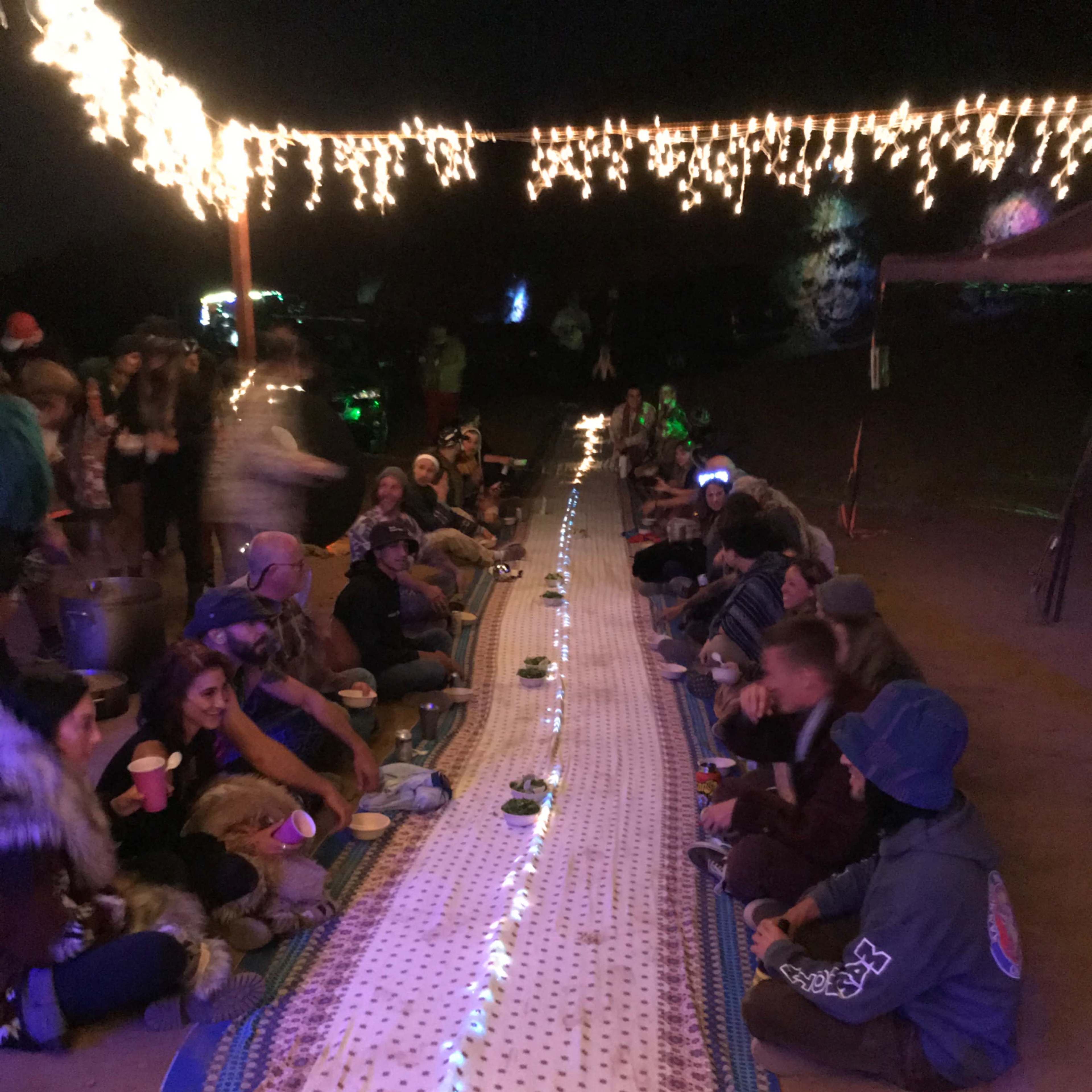 A long, illuminated table covered with a patterned cloth is set outdoors, surrounded by a group of people sitting on the ground while holding cups.