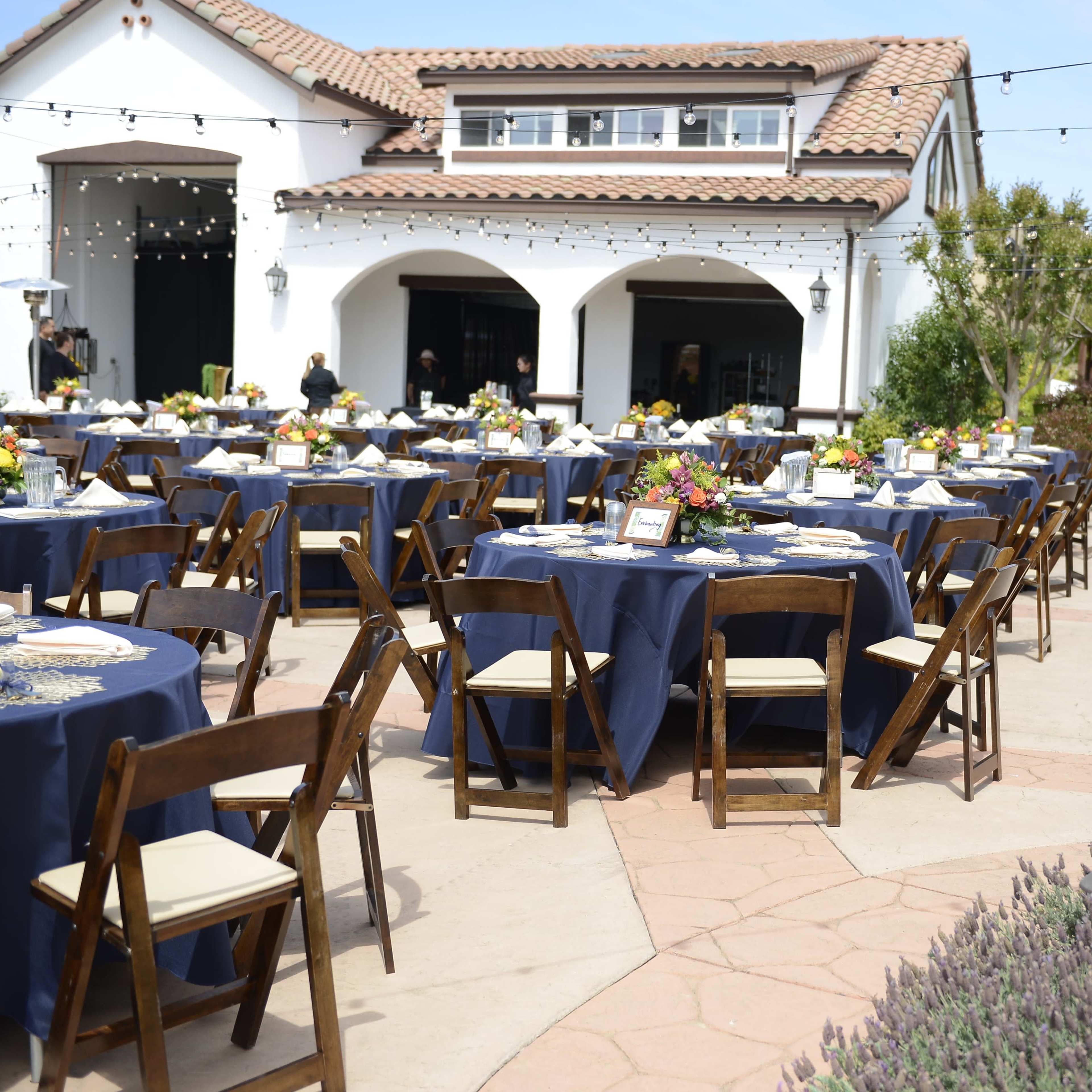 An outdoor dining setup with round tables covered in navy blue tablecloths arranged neatly in front of a white building, surrounded by greenery and string lights.