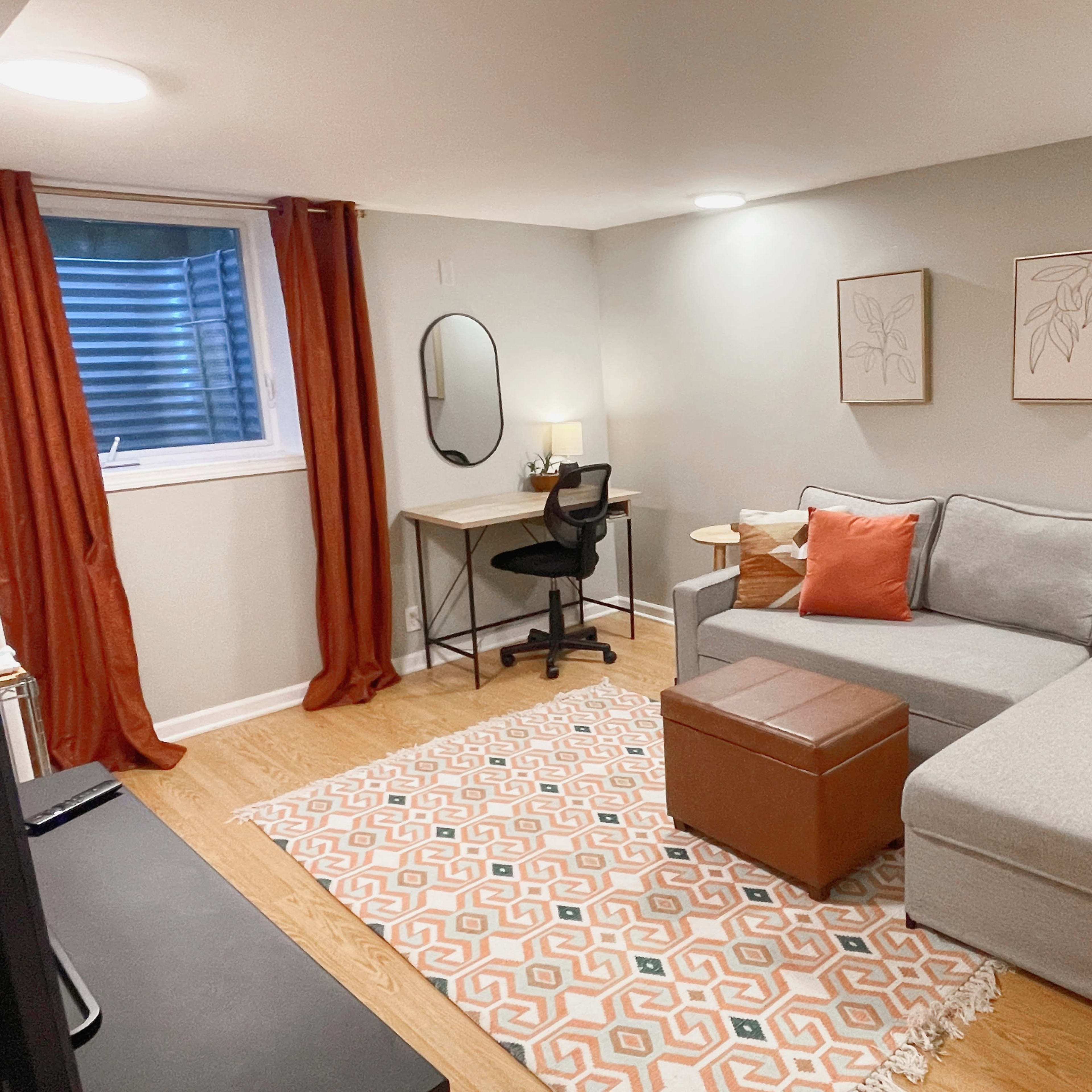A cozy living area featuring a gray sectional couch, a small desk with a chair, and decorative wall art, all set against a light-colored backdrop with orange curtains.