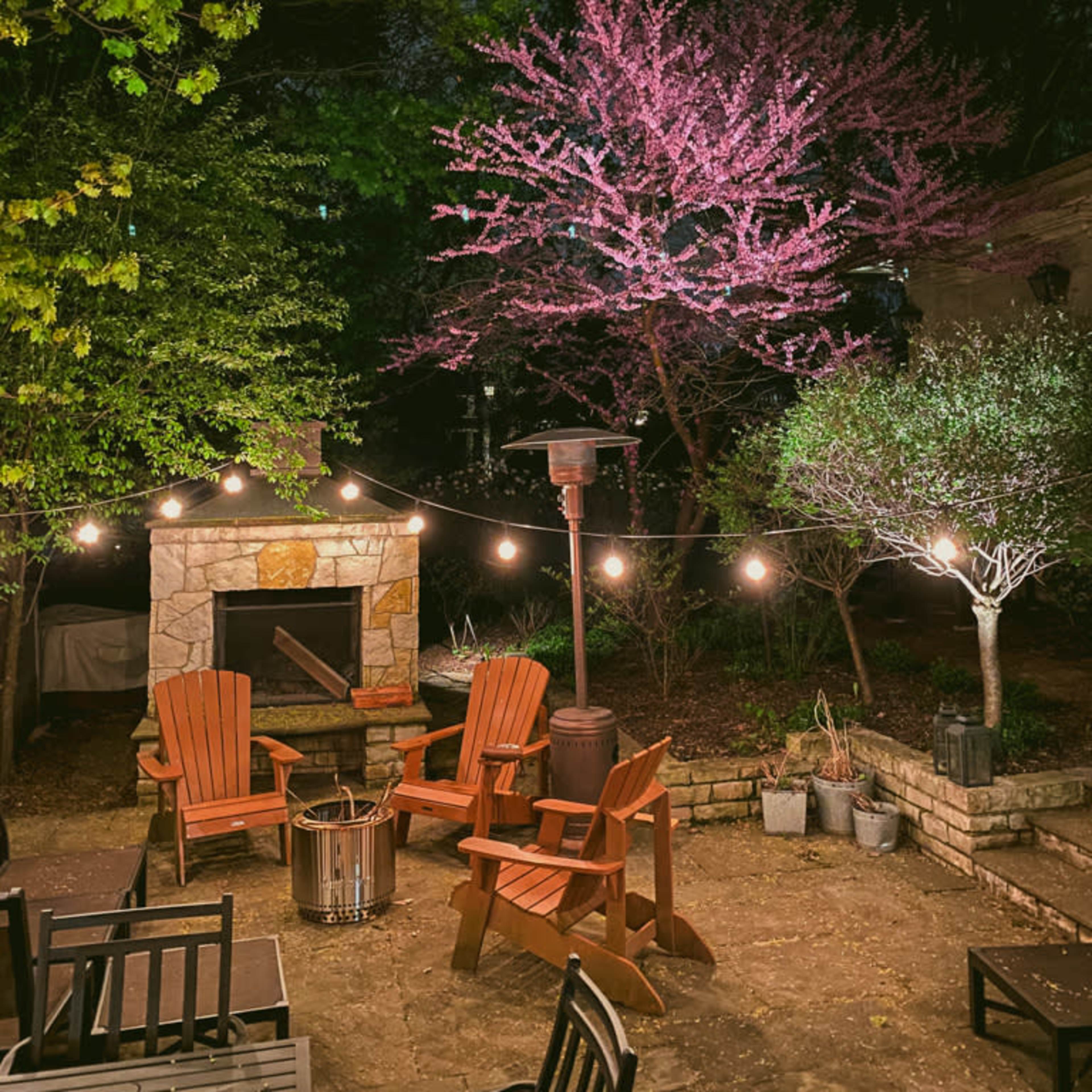 A landscaped backyard at night, featuring an outdoor seating area with wooden chairs, a stone fireplace, and a flowering tree illuminated by string lights.