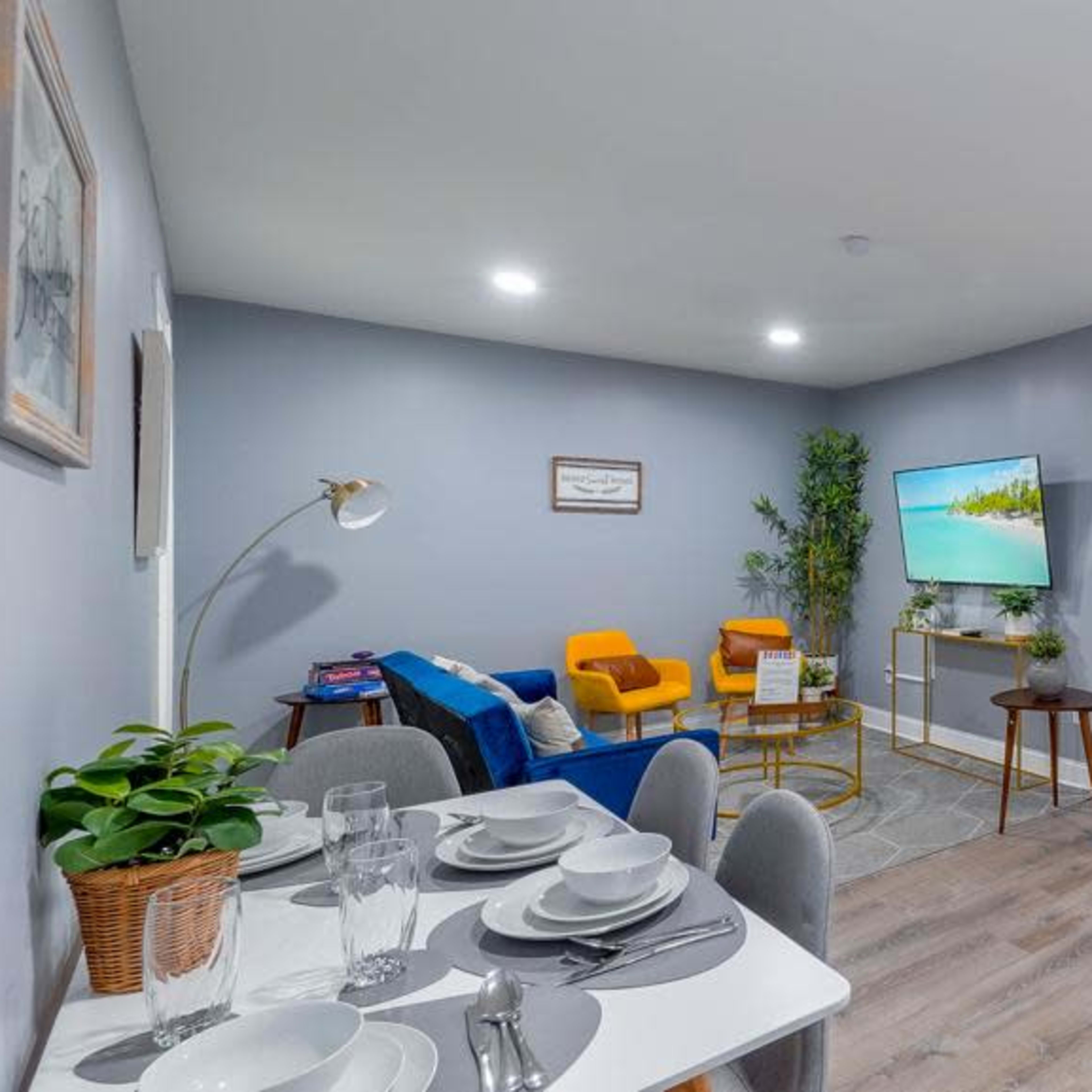 A modern open-concept living space features a dining area with a table set for four, a seating area with colorful chairs, a television mounted on the wall, and a visible bedroom through an archway.