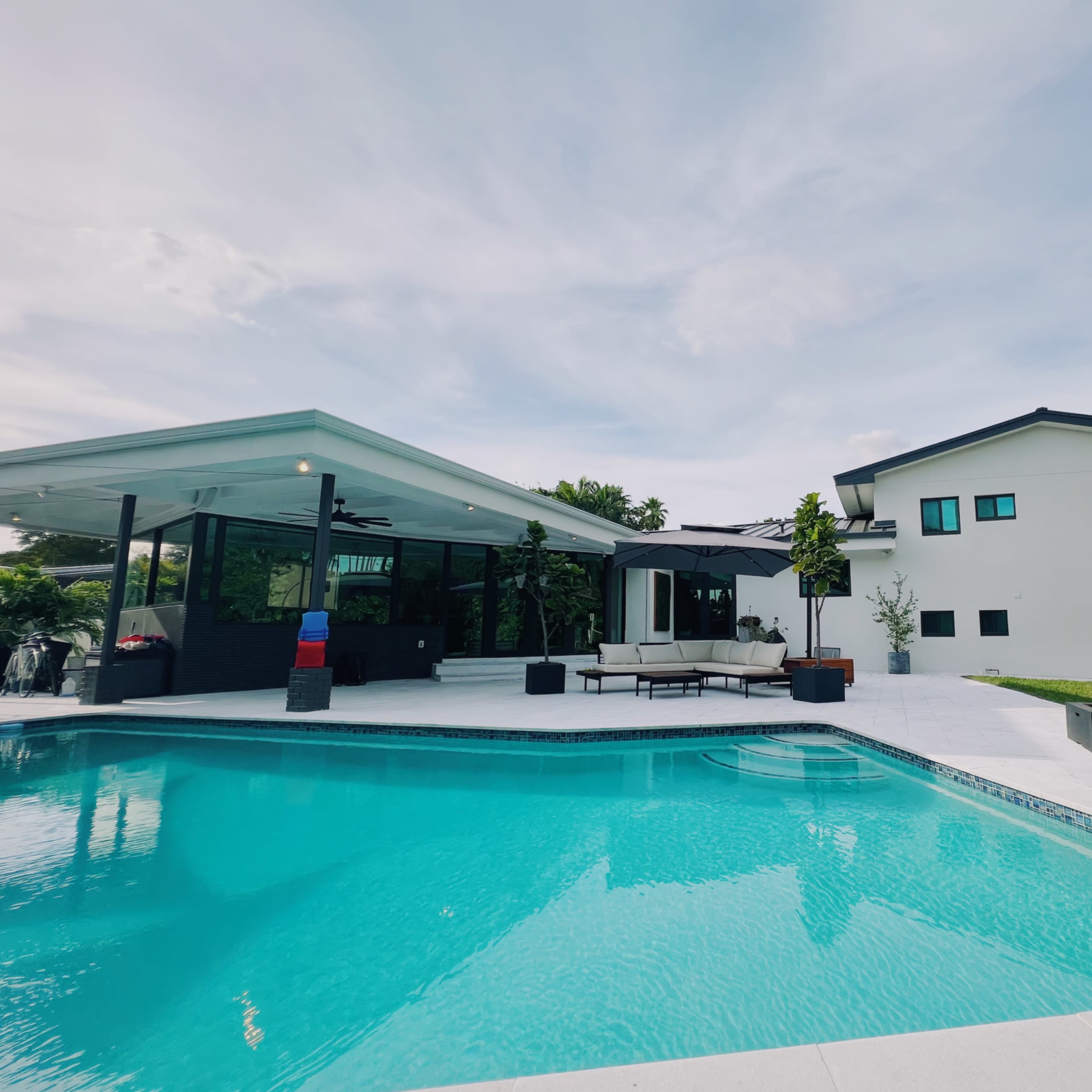 A modern house with a large swimming pool and patio area is situated in a green yard under a blue sky.