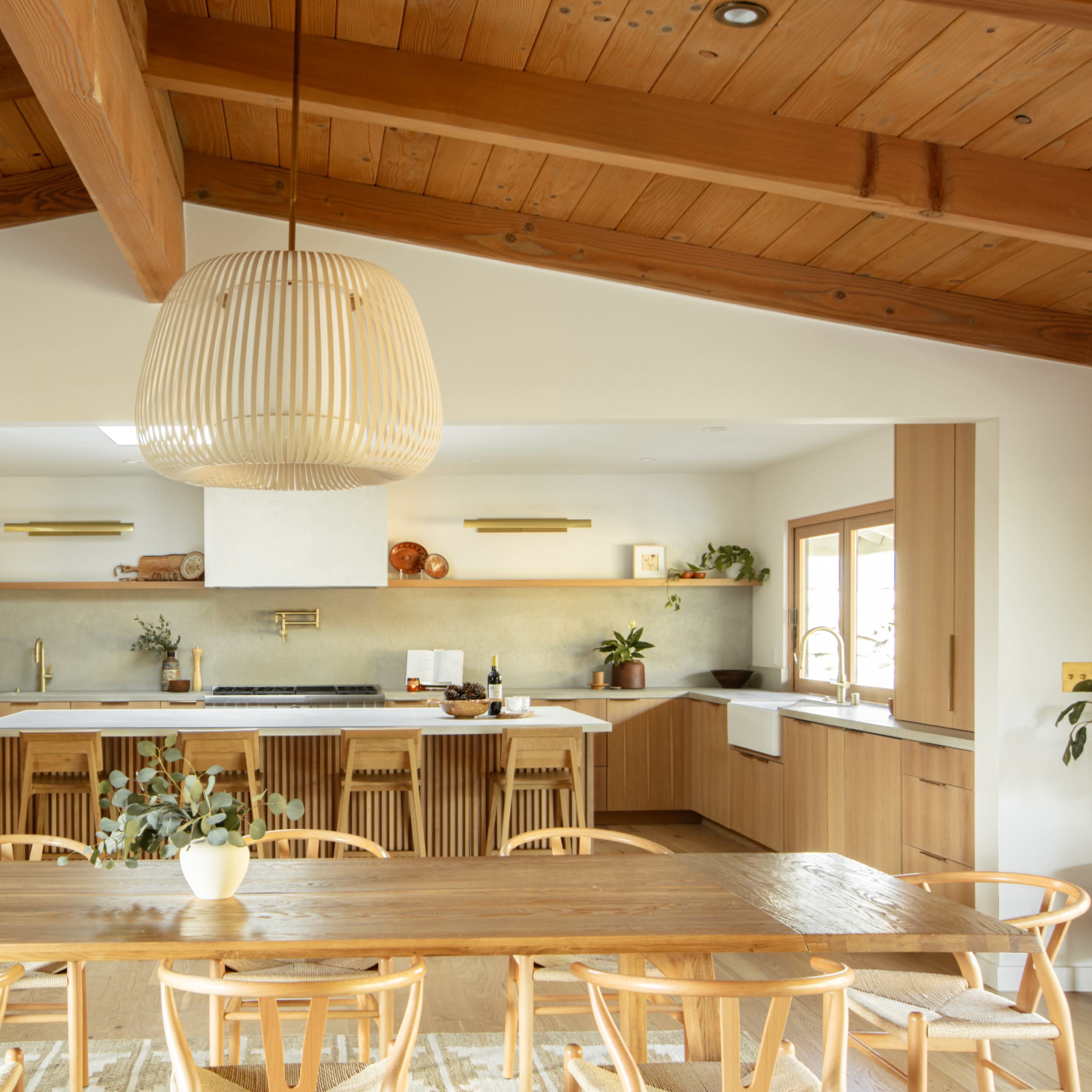 A modern kitchen and dining area with a long wooden table, a sloped wooden ceiling, and contemporary cabinetry.