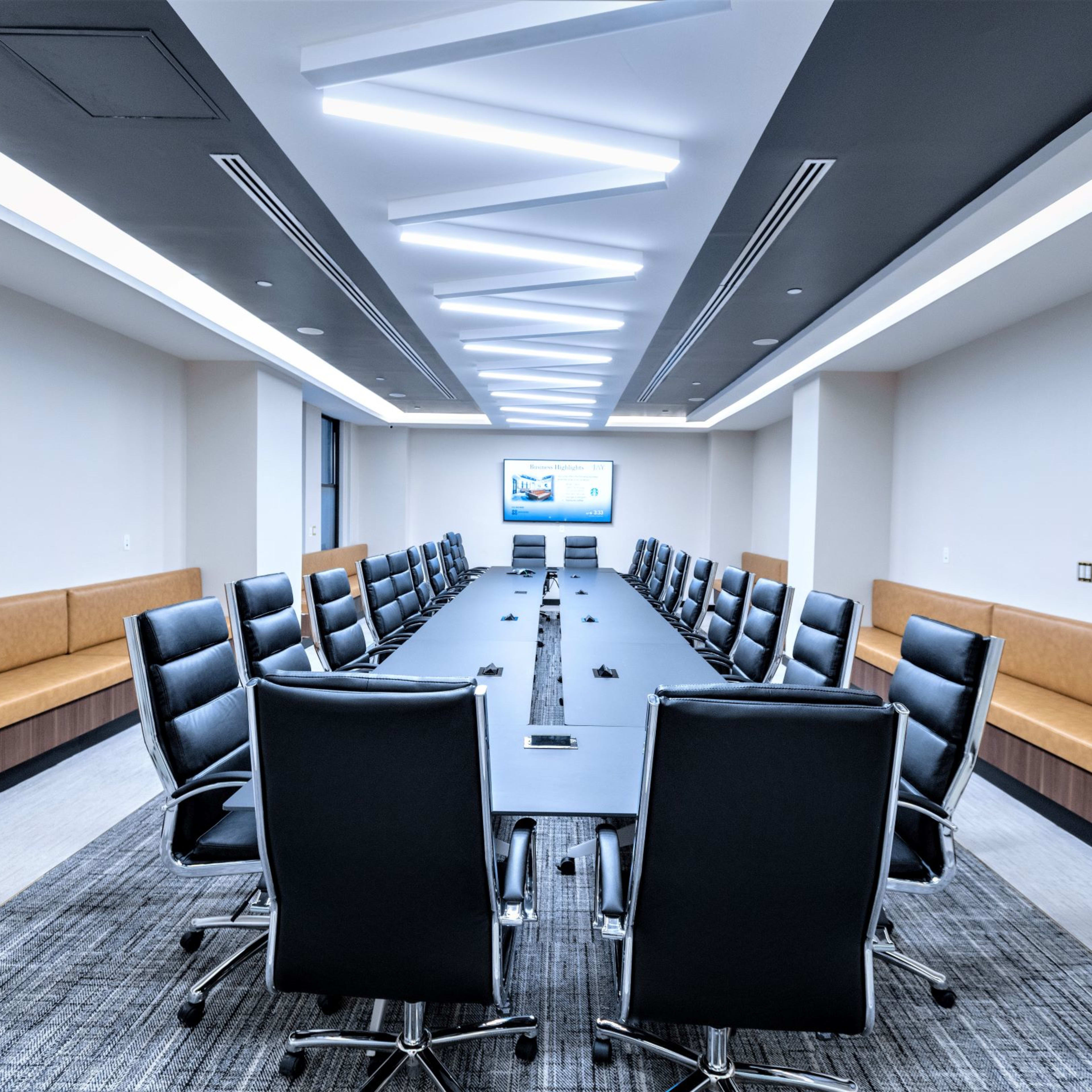 A modern conference room features a long table surrounded by ergonomic black chairs and a presentation screen at one end, illuminated by linear ceiling lights.