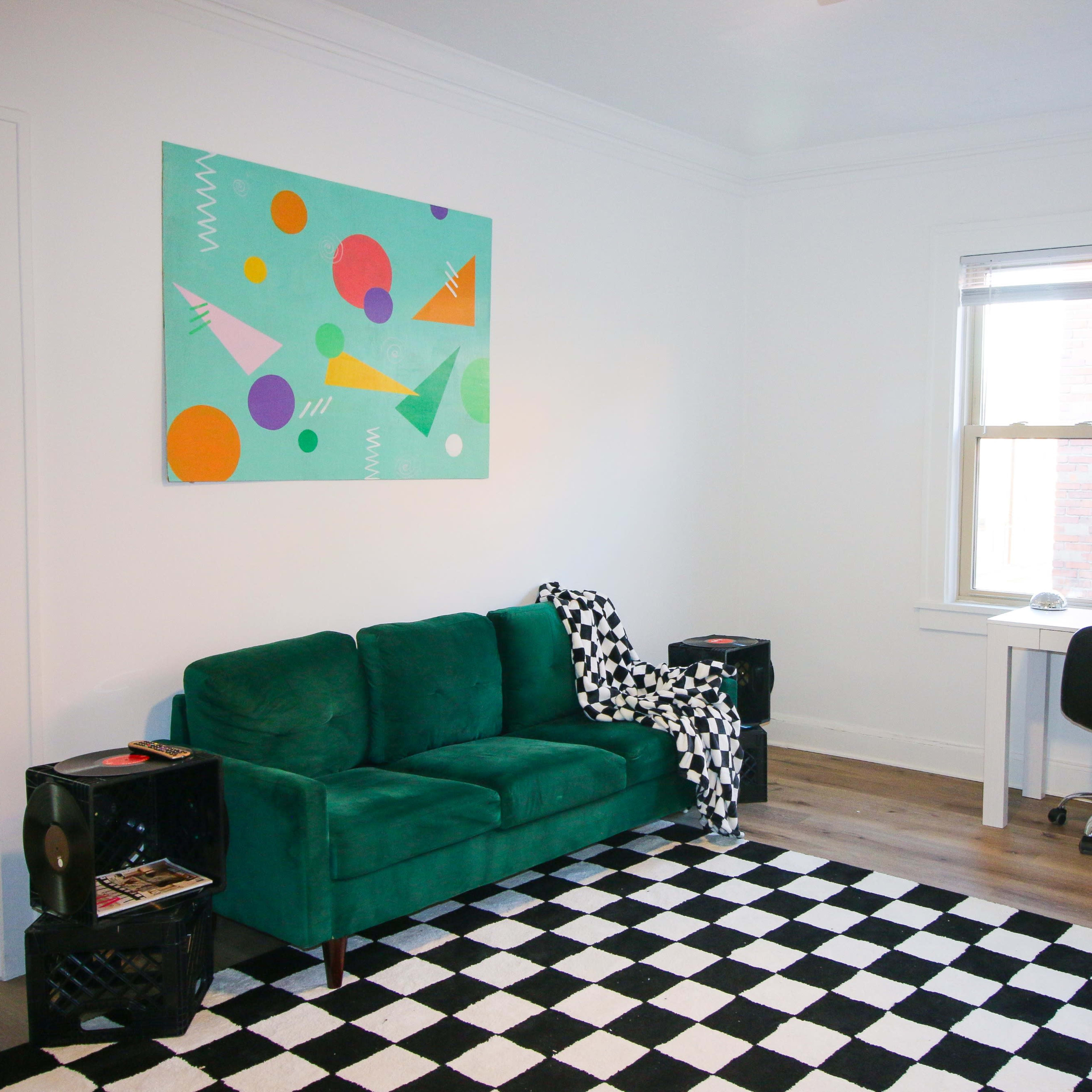 The room features a green sofa, a black and white checkered rug, a white desk with a chair, and an abstract painting on the wall.