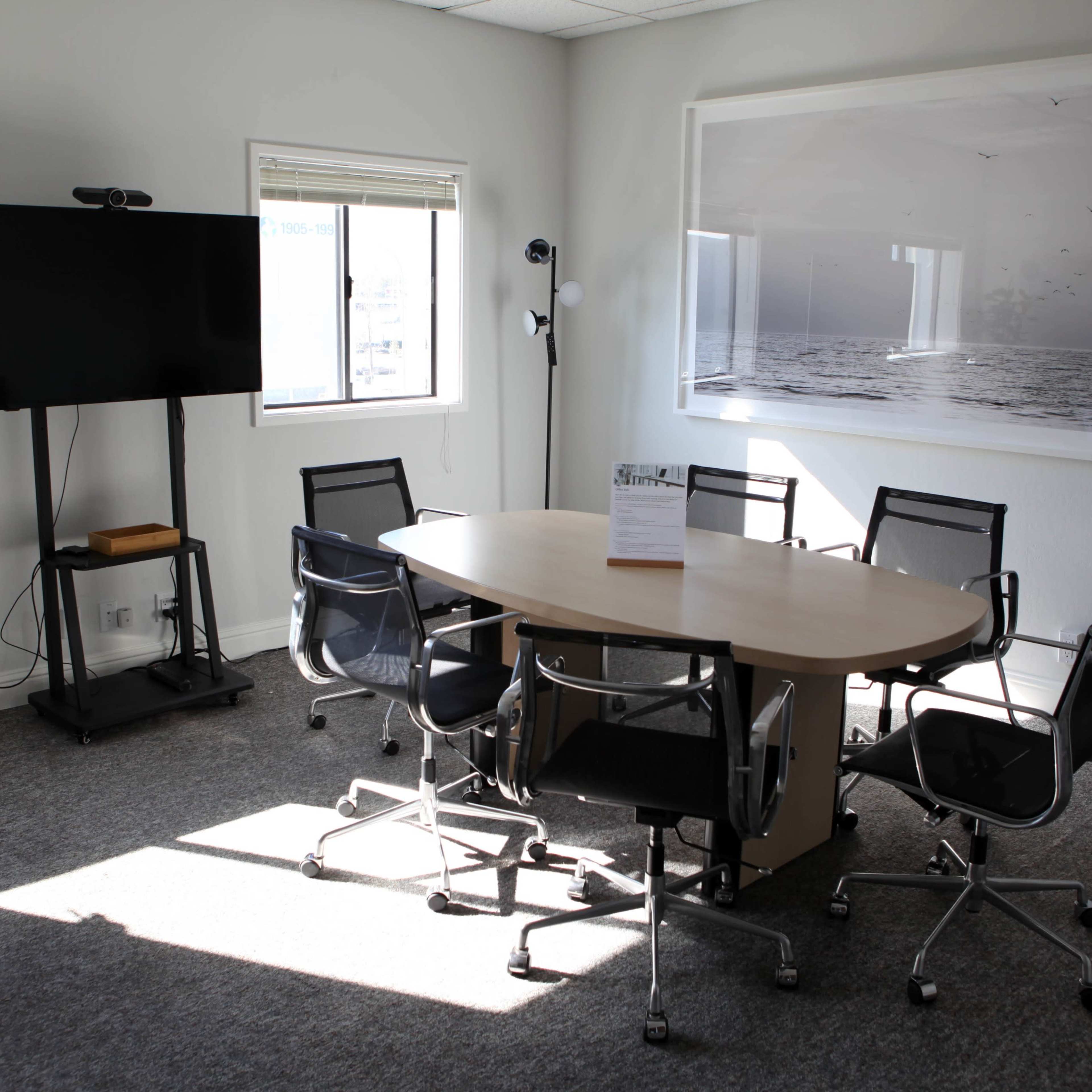 A conference room features a round wooden table surrounded by six black chairs, a television on a stand, and large windows with a view.
