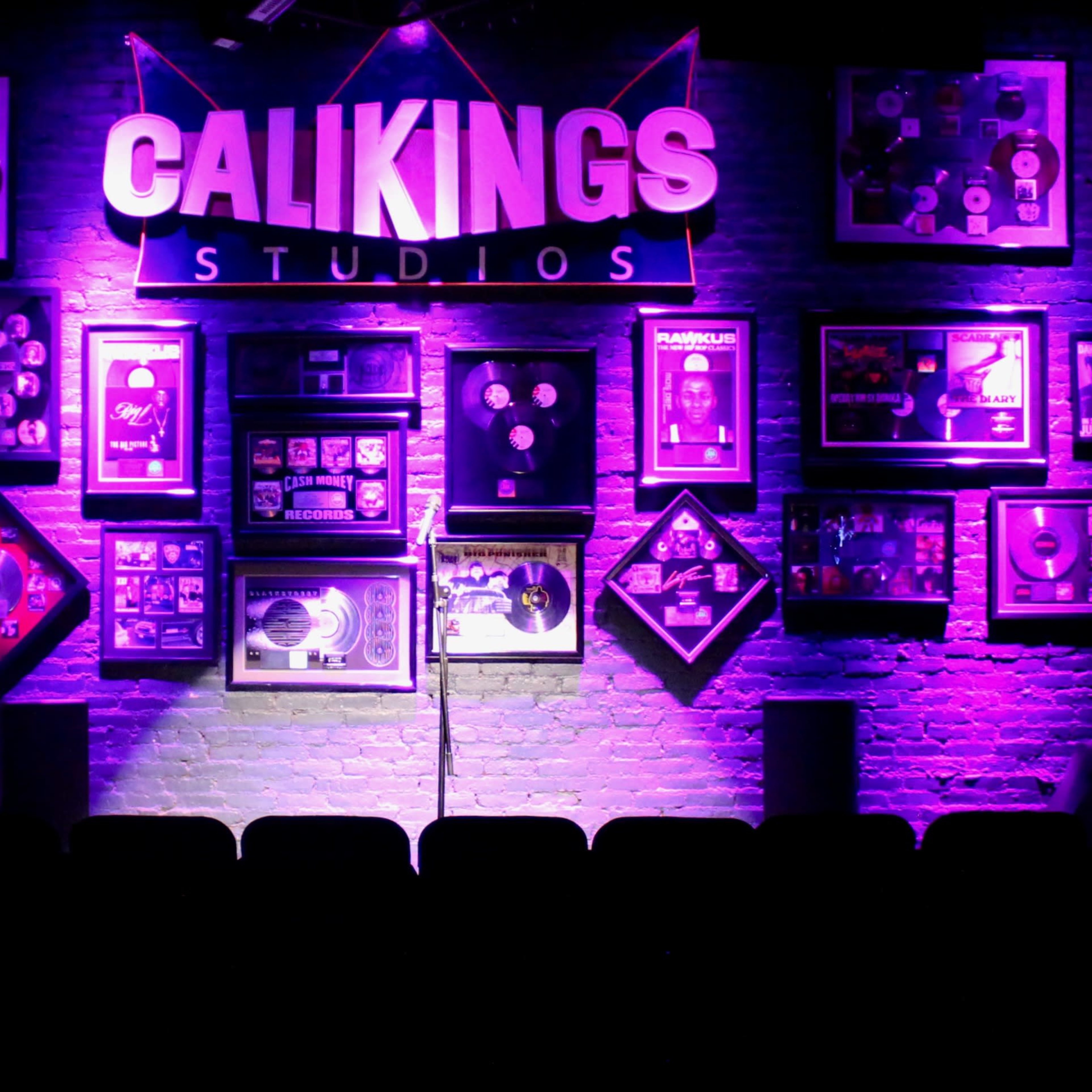A performance stage with a microphone in front of a brightly lit wall displaying framed records and awards at Calikings Studios.