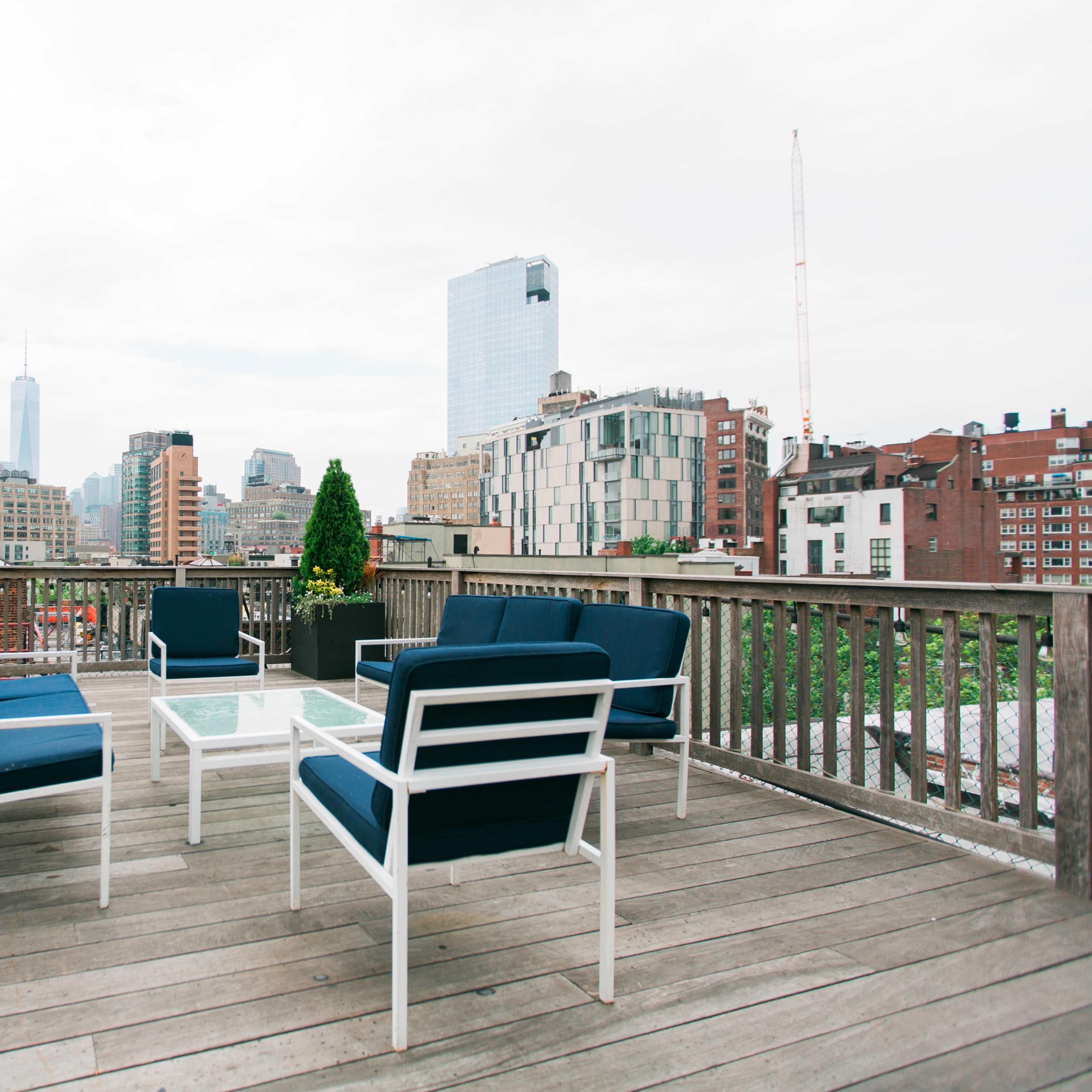 A rooftop terrace with modern furniture overlooks a city skyline featuring tall buildings and a cloudy sky.