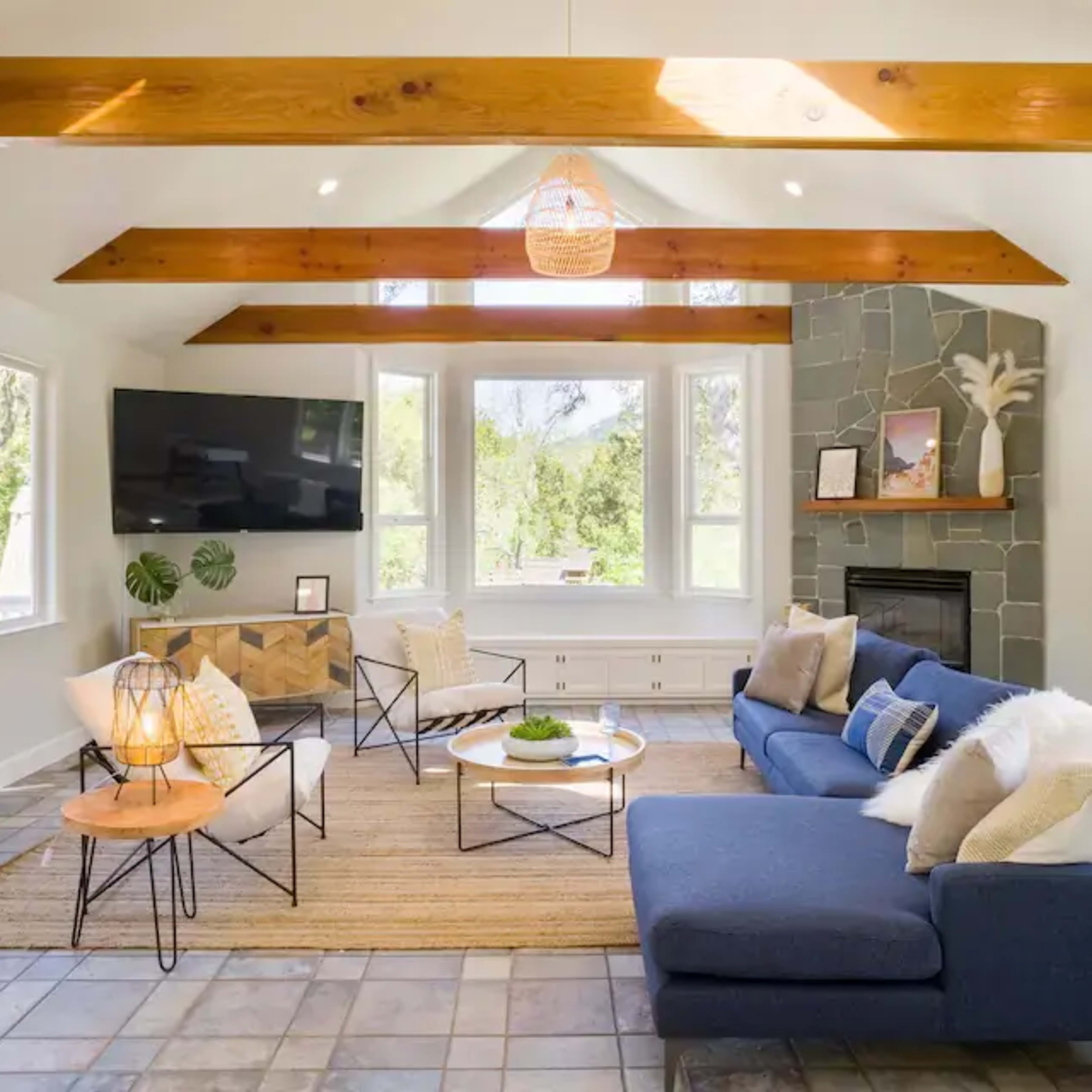 The living room features a blue sofa, a television mounted on the wall, and large windows that provide a view of the outdoors, all under wooden beam ceilings.