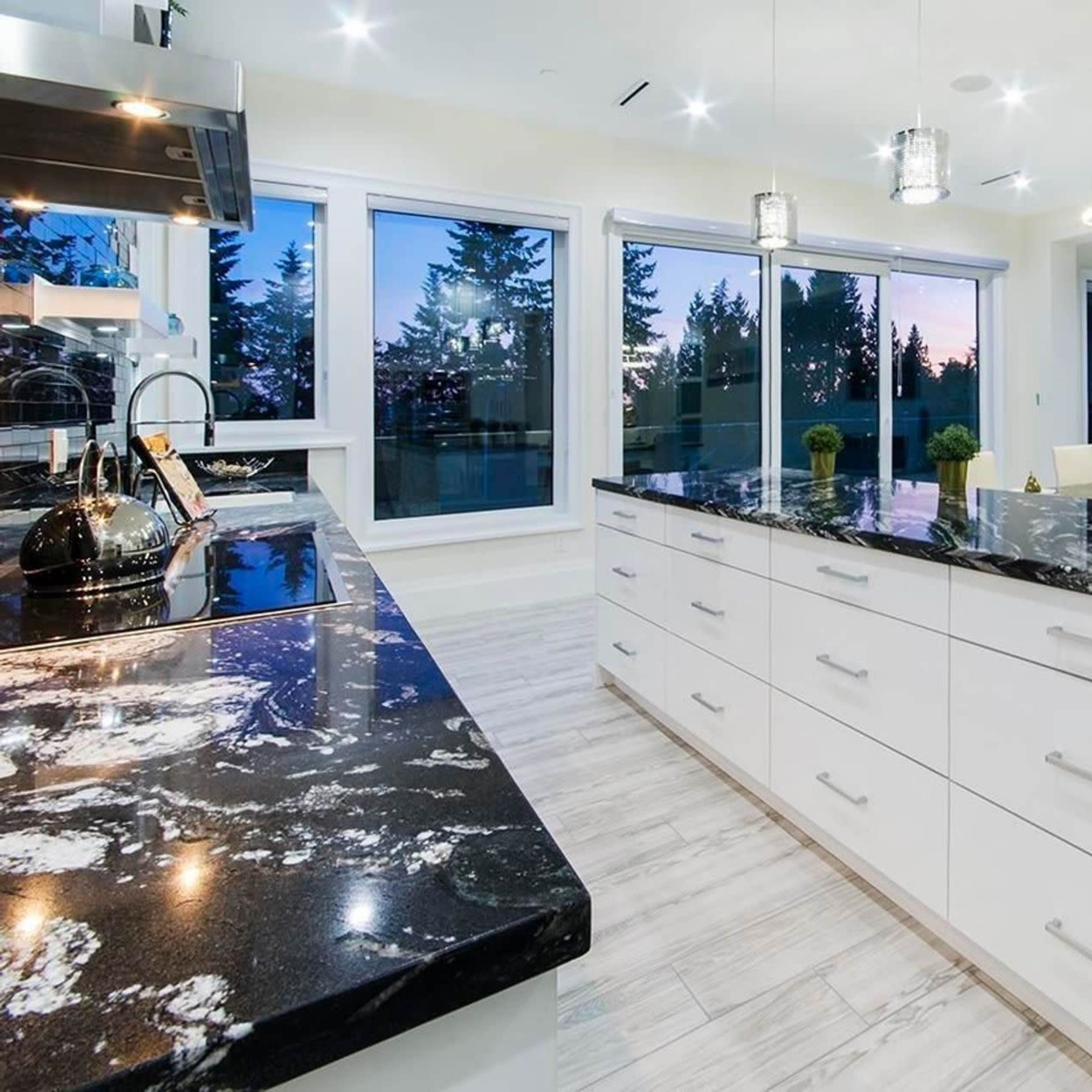 A modern kitchen features a large island with a dark marble countertop and white cabinetry, overlooking a view of trees and a sunset through large windows.