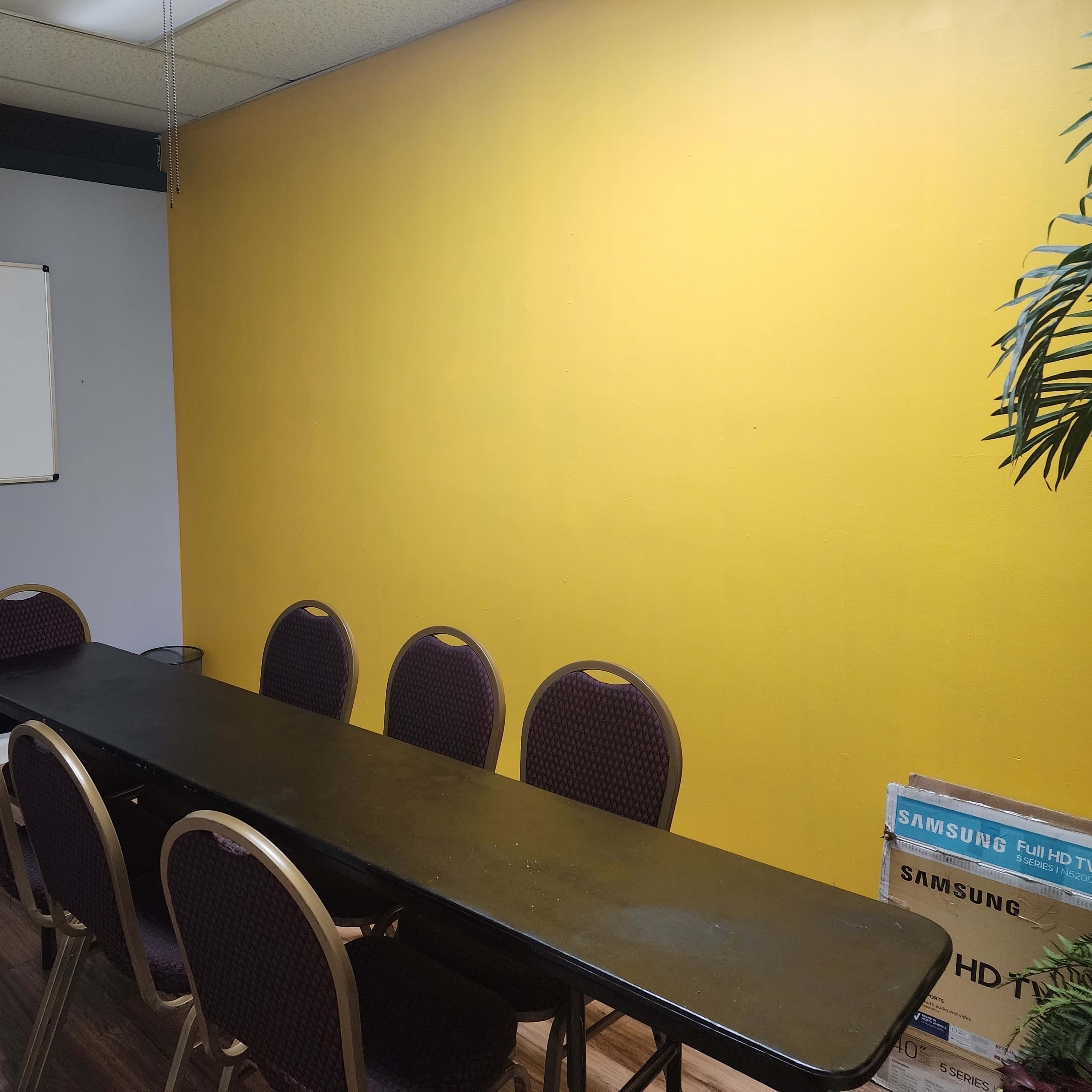 A meeting room with a long black table and several purple-upholstered chairs, adjacent to a yellow wall and a whiteboard.