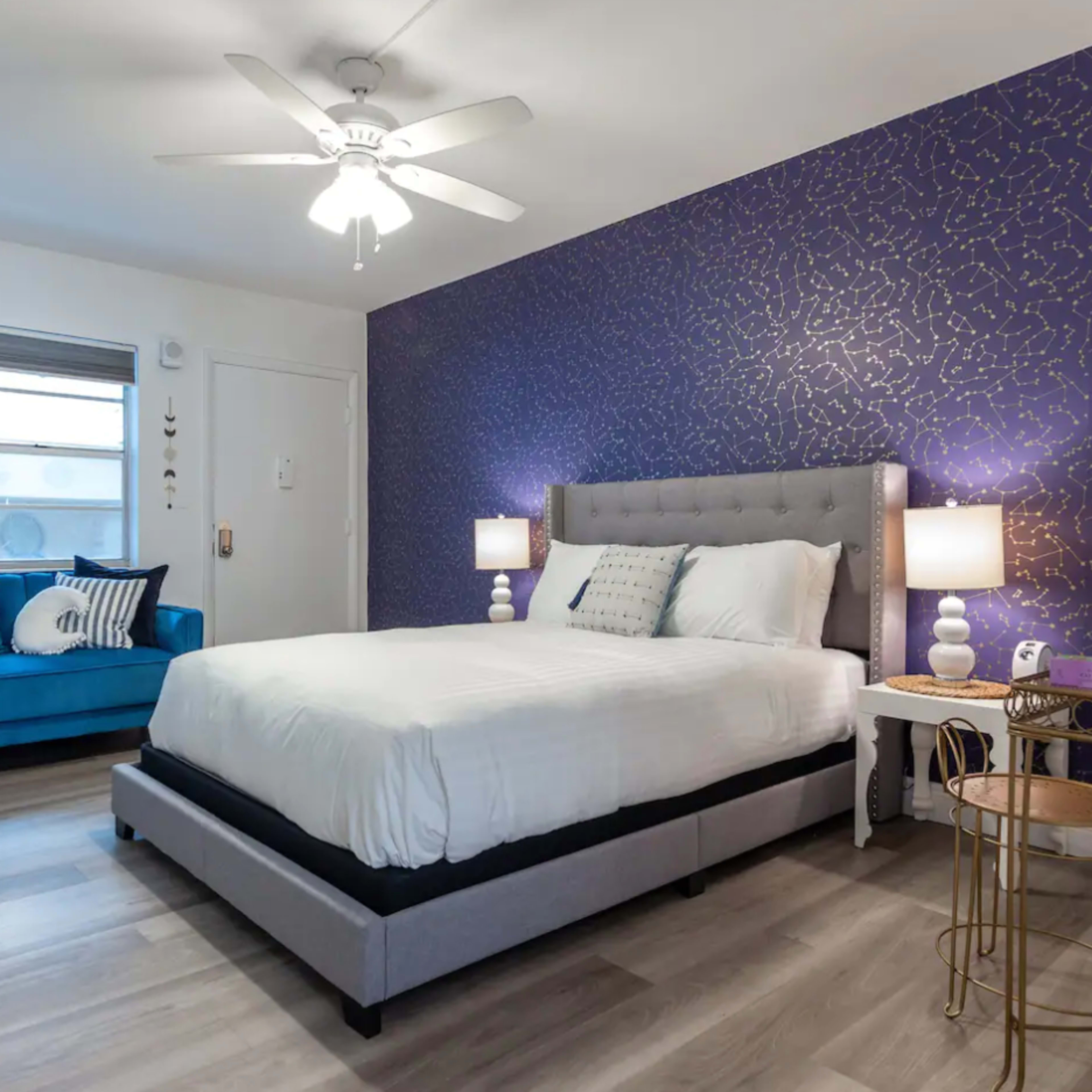 A compact bedroom featuring a light-colored bed, a teal sofa, and a purple accent wall with a textured design.