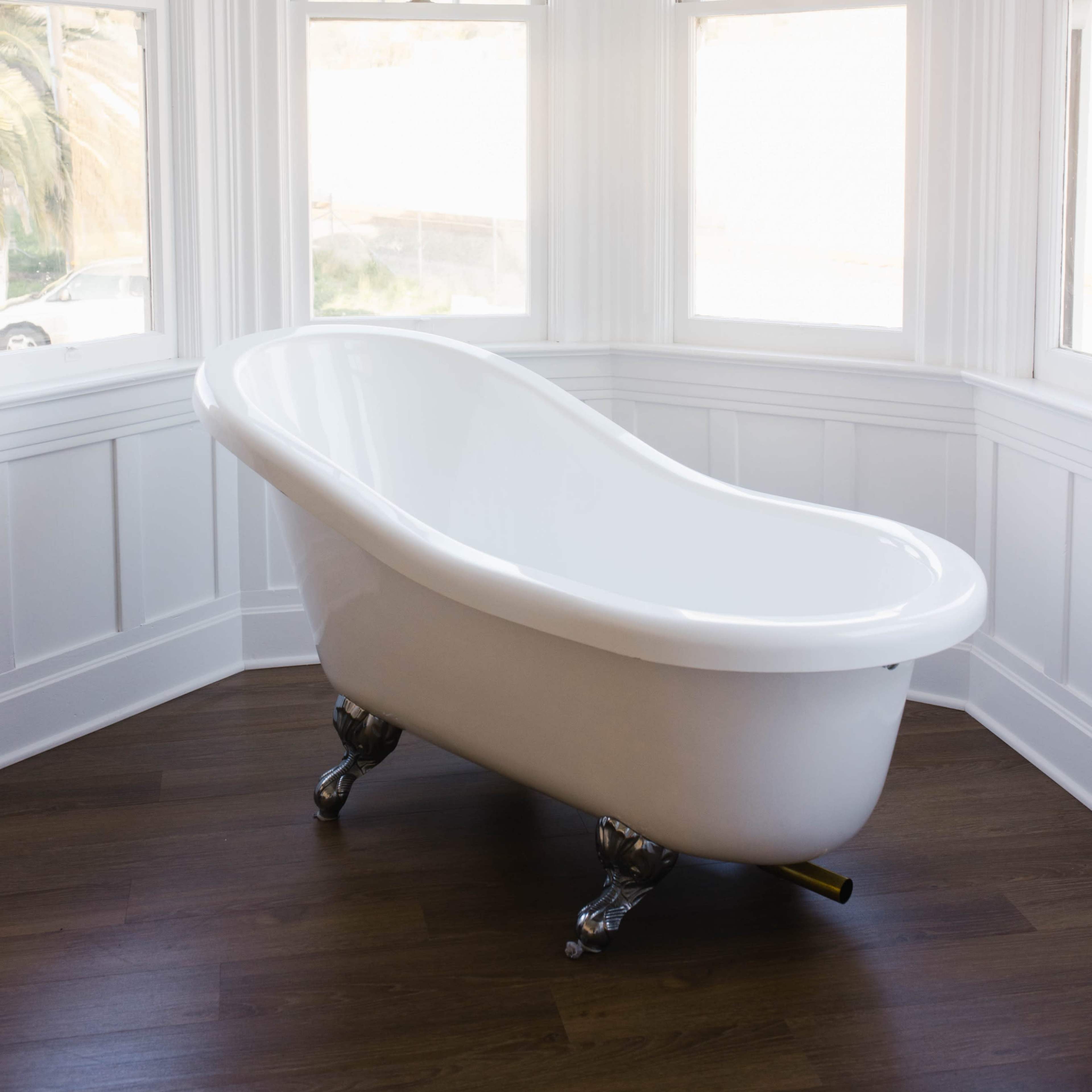 A white, freestanding bathtub is positioned in a corner of a room with large windows on two sides and wooden flooring.