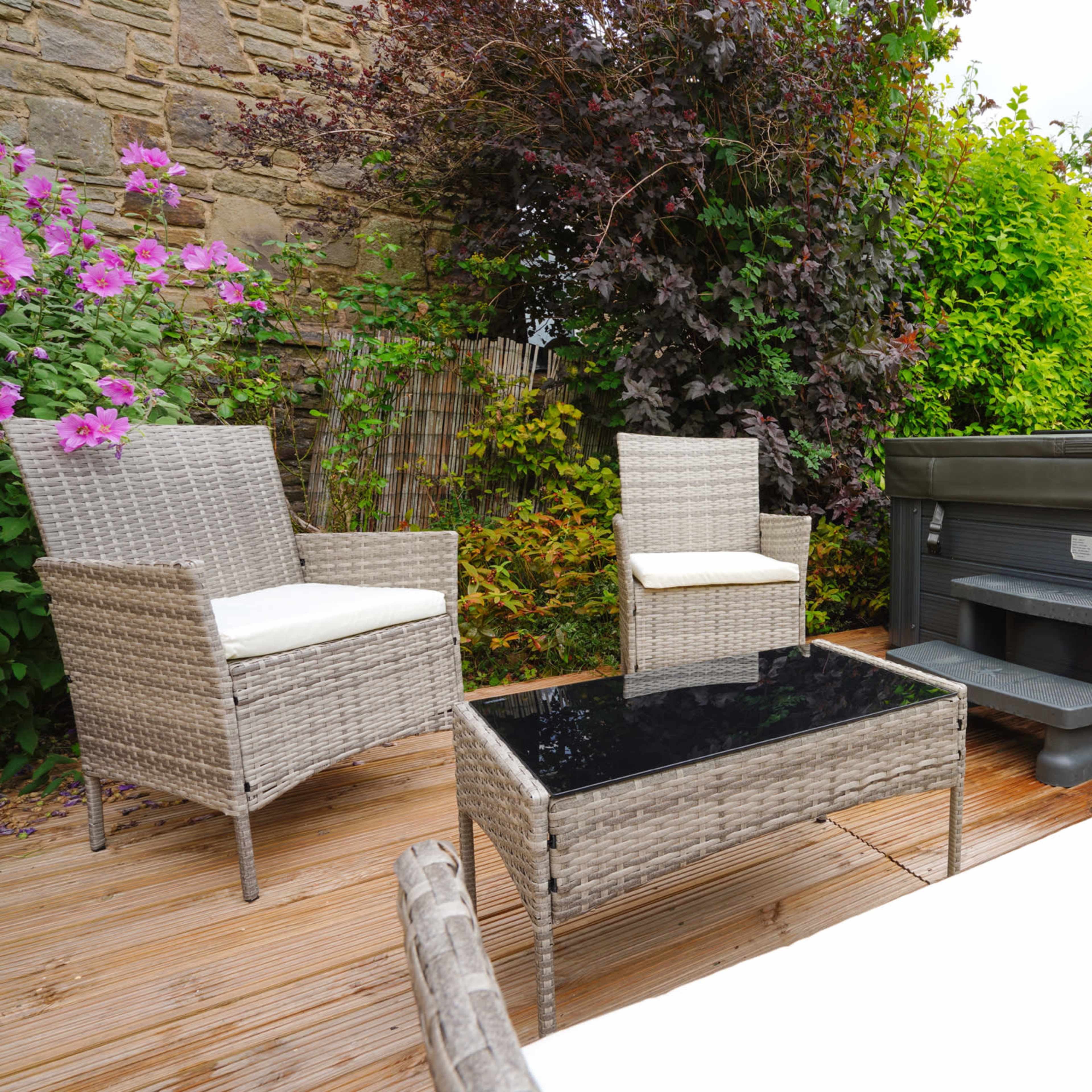 A patio area features two wicker chairs and a glass-top table beside a hot tub, surrounded by lush greenery and flowers.
