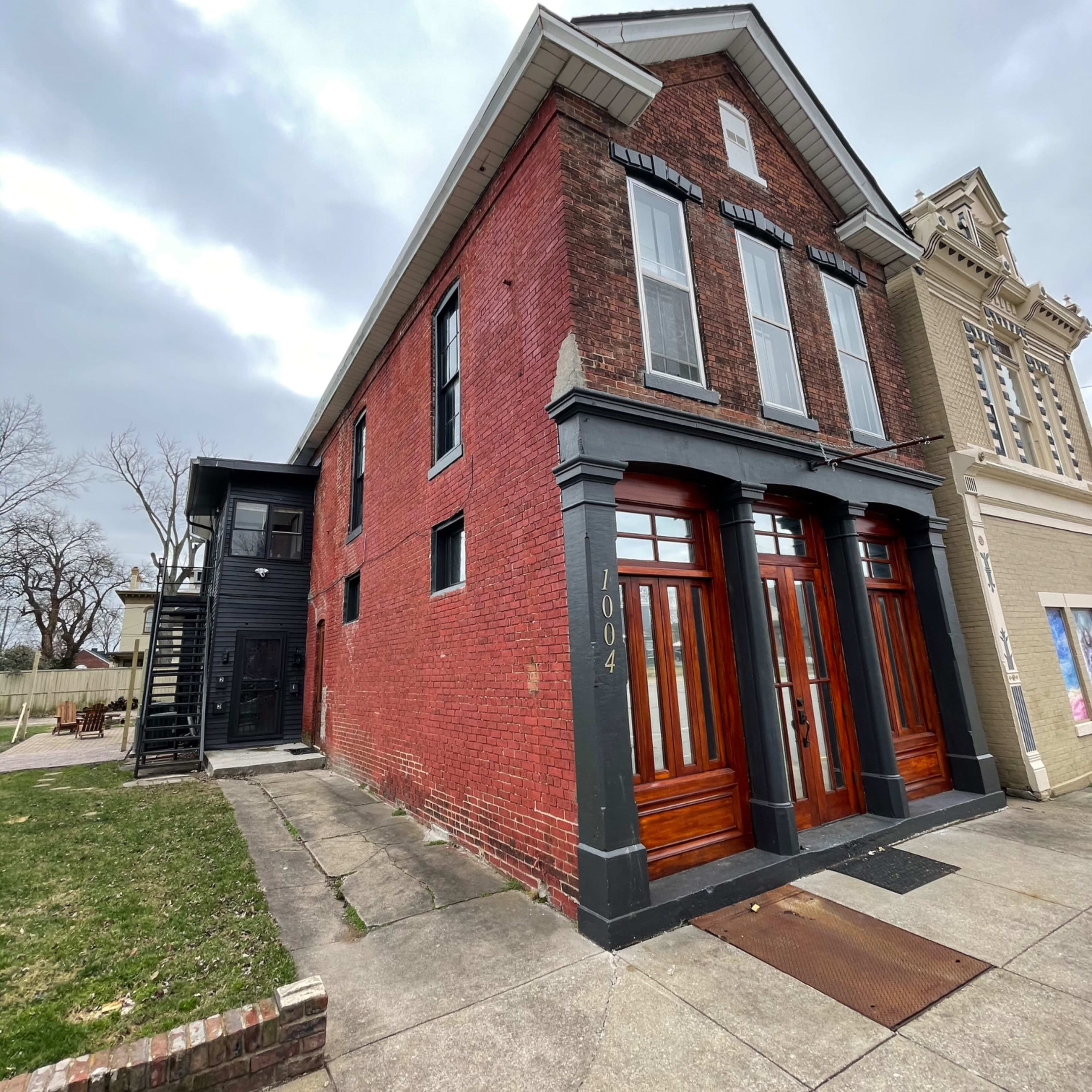 A two-story red brick building with large wooden doors, situated on a corner lot adjacent to another building.