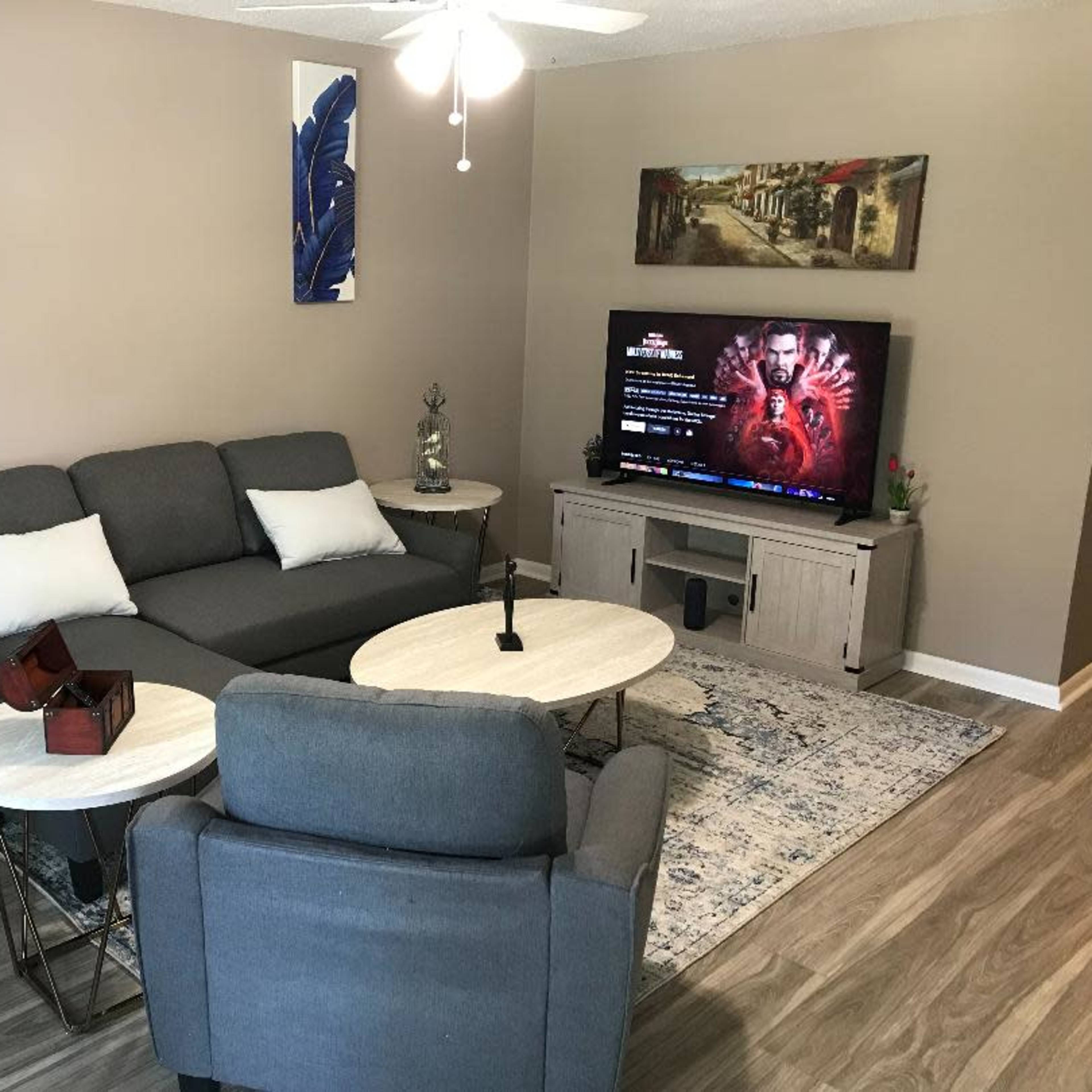 A living room with a gray sectional sofa, two small coffee tables, a television displaying content, and decorative wall art.