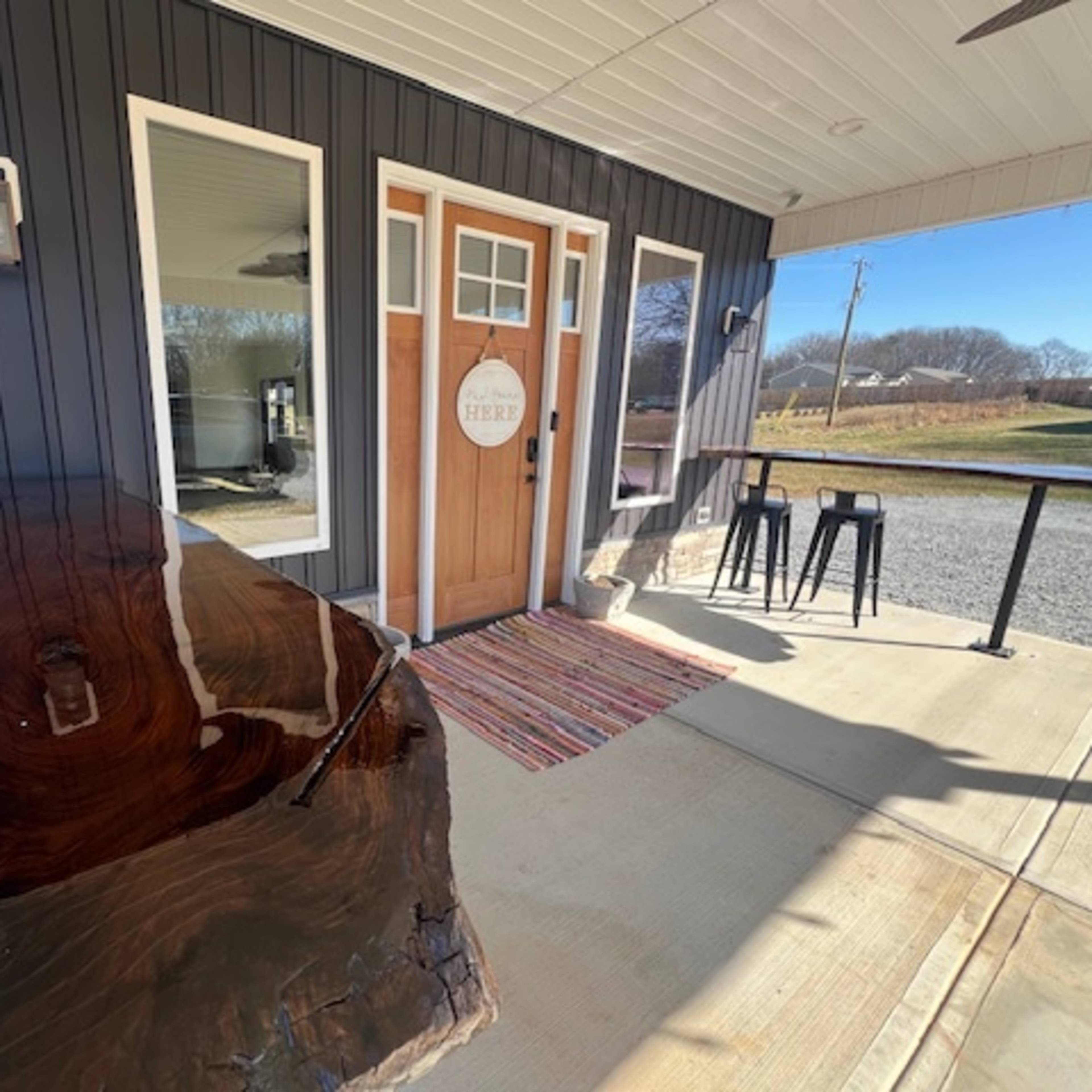 A wooden table is positioned beside the entrance of a modern building with a sign that reads "HERE," featuring large windows and a porch area.