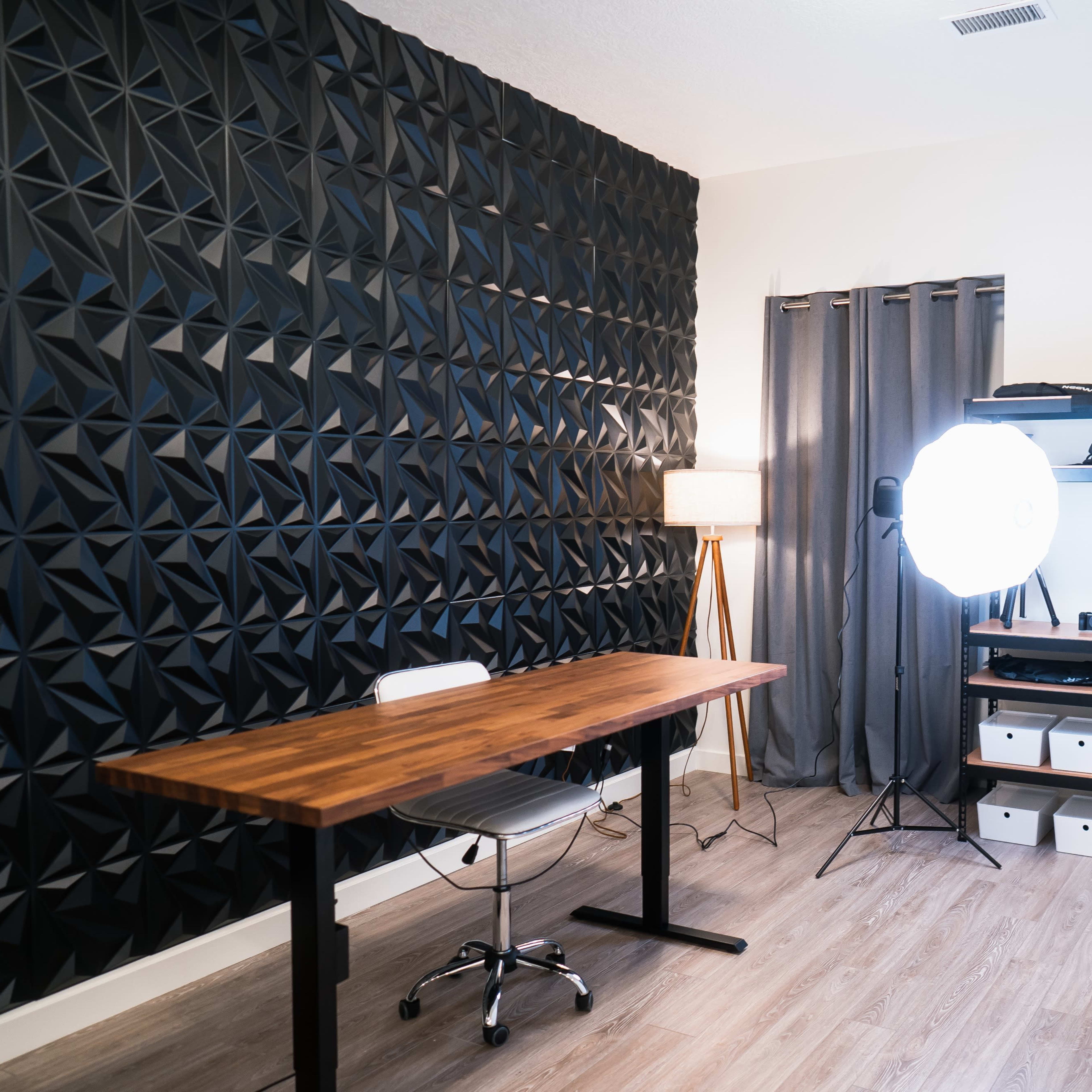 A minimalist office space featuring a wooden desk, a large light source, storage boxes, and a textured black wall.