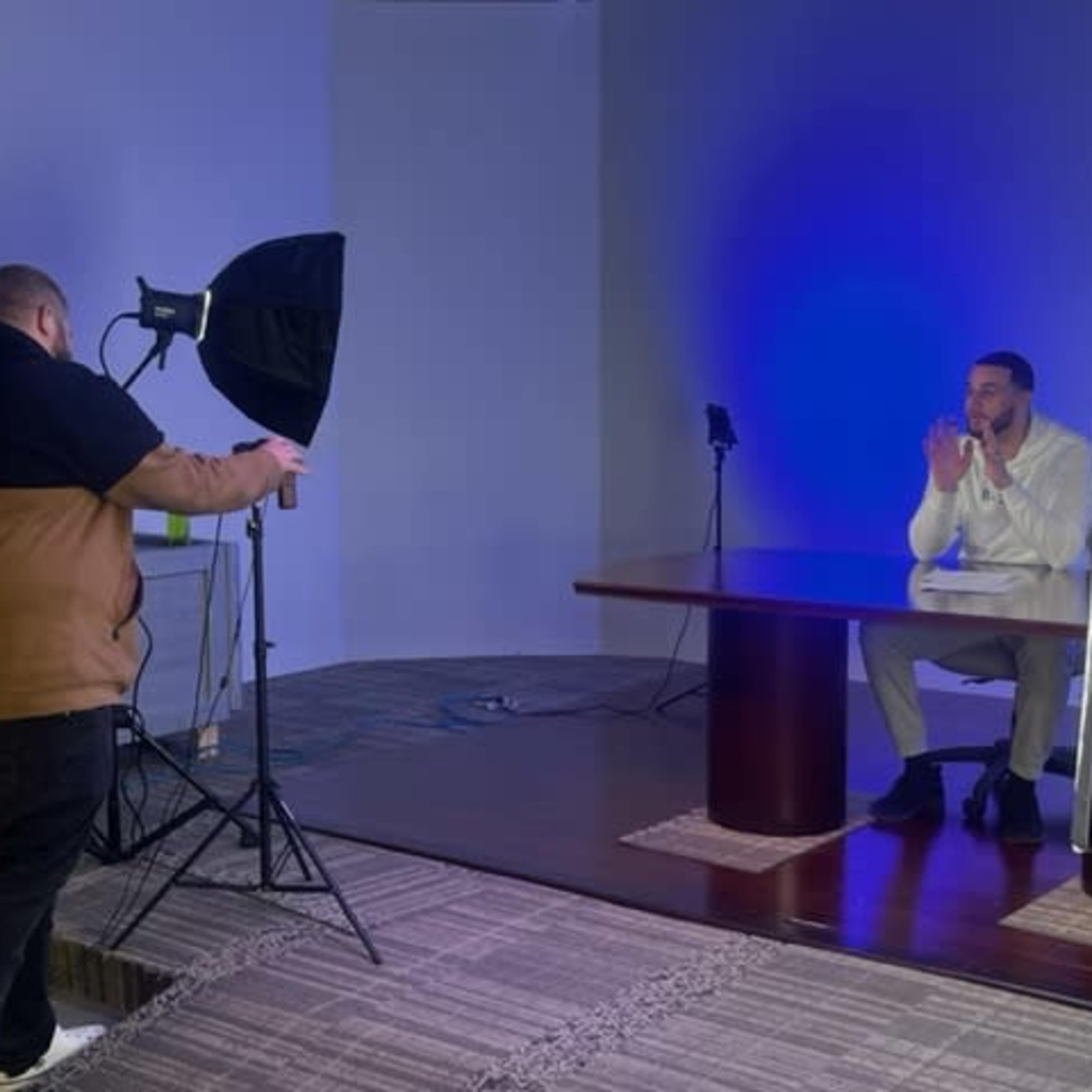 A man sits at a table in a studio while another man adjusts a light setup and camera equipment.