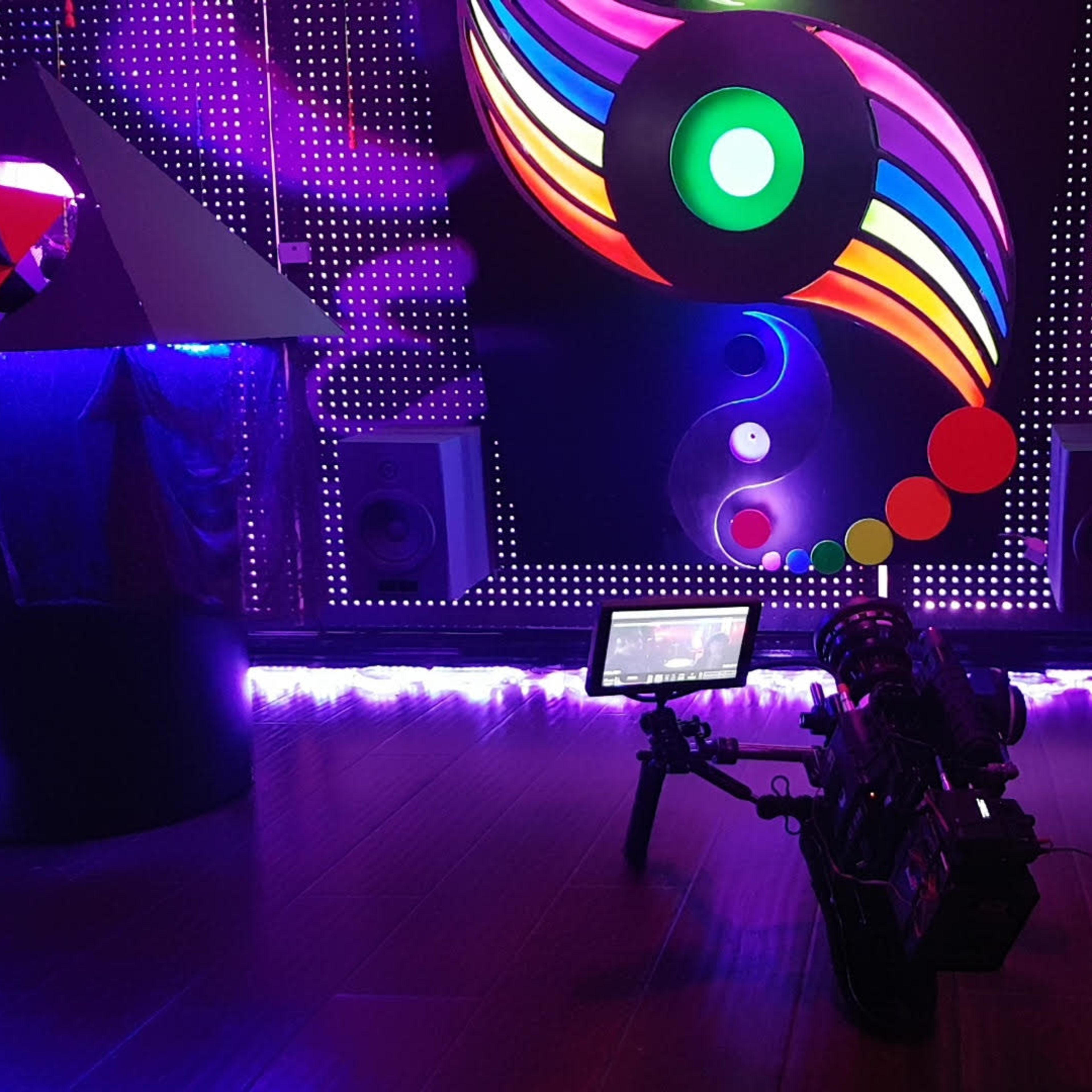 The scene features a colorful illuminated backdrop with circular patterns, a pyramid sculpture, speakers, and a camera set up on a wooden floor.