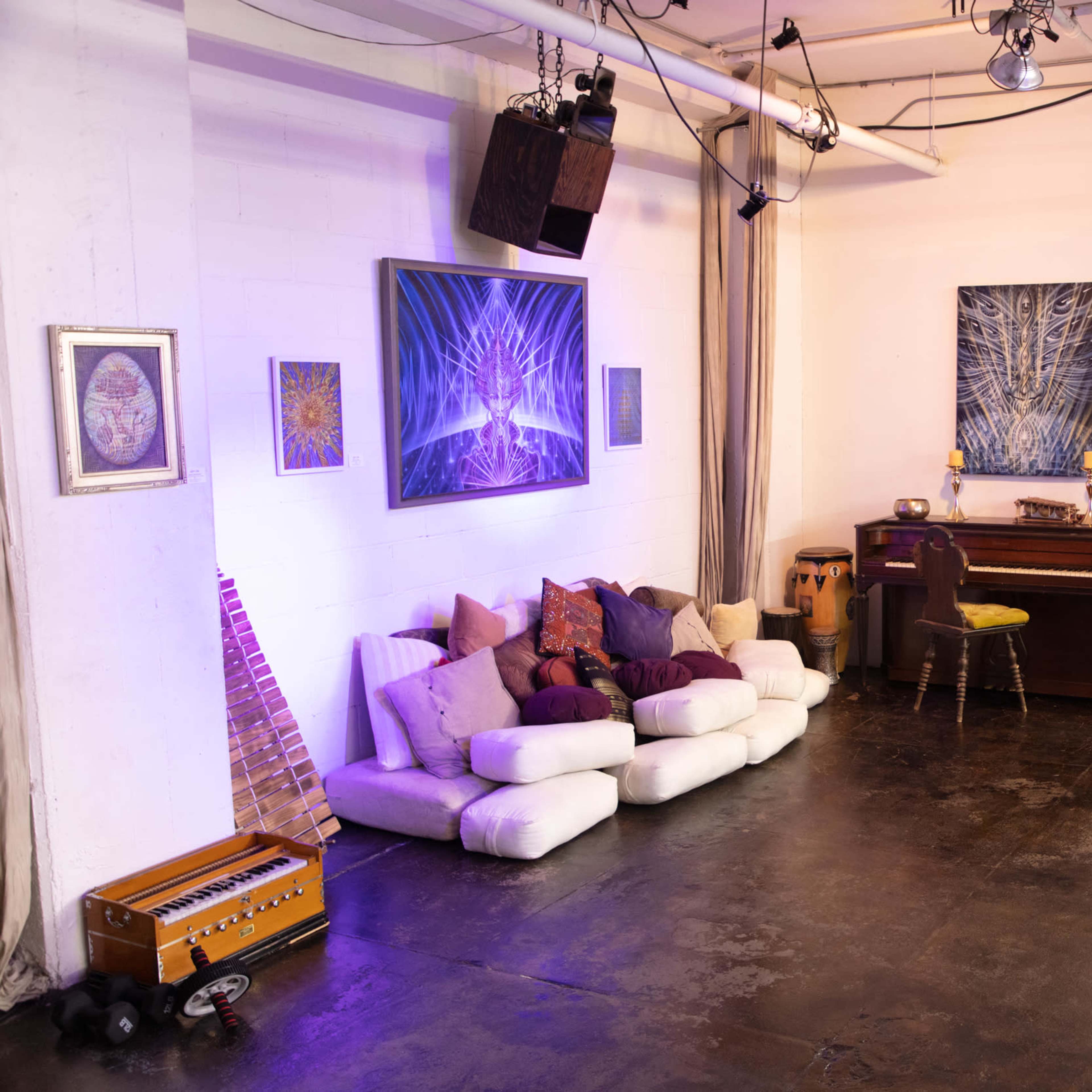 A spacious studio with a seating area on the left, a piano in the center, and various artworks hanging on the walls.