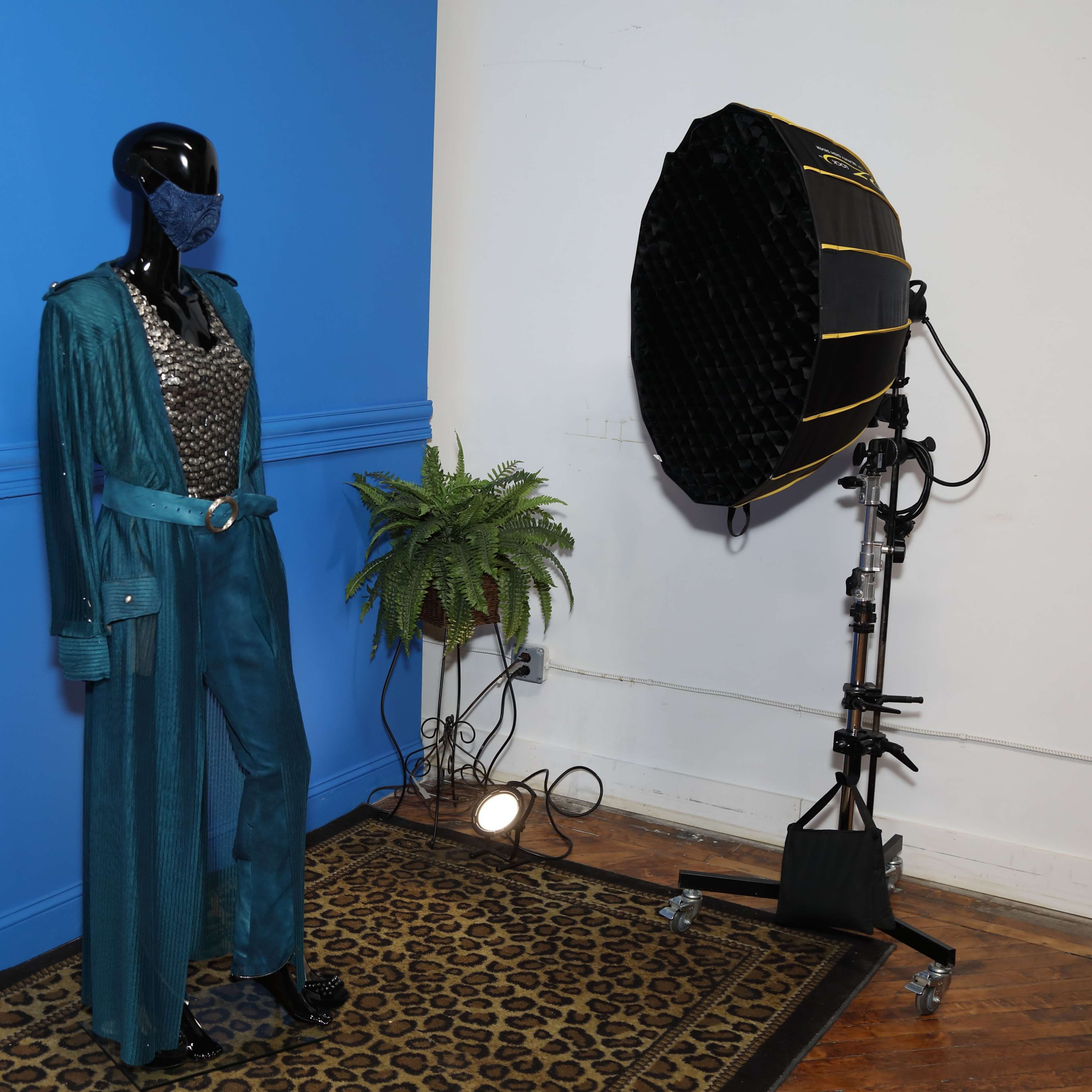A mannequin wearing a teal outfit stands next to a large softbox light in front of a blue wall and a patterned rug.