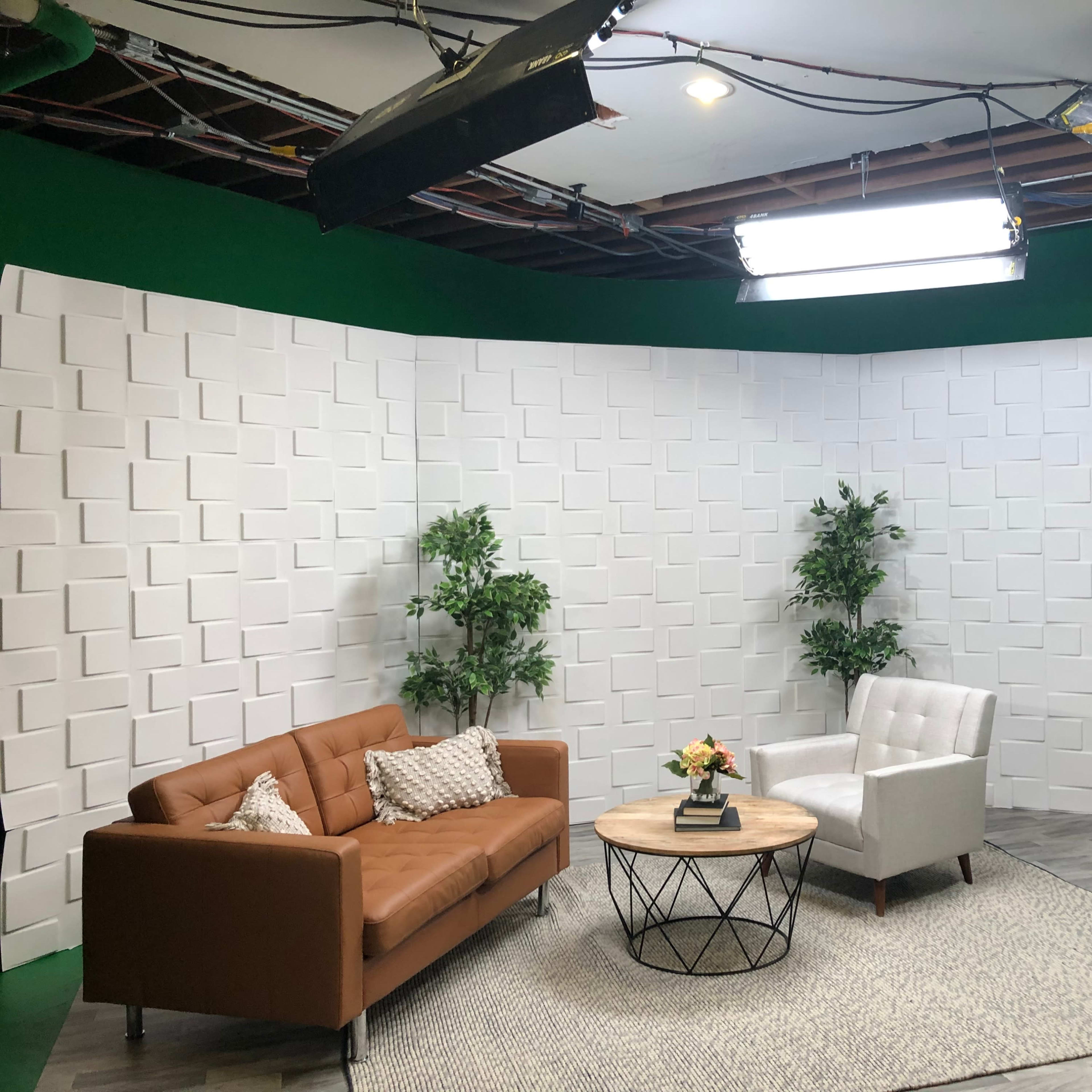 A TV studio set with a brown sofa, a white armchair, a round coffee table, two plants, and a textured white backdrop.