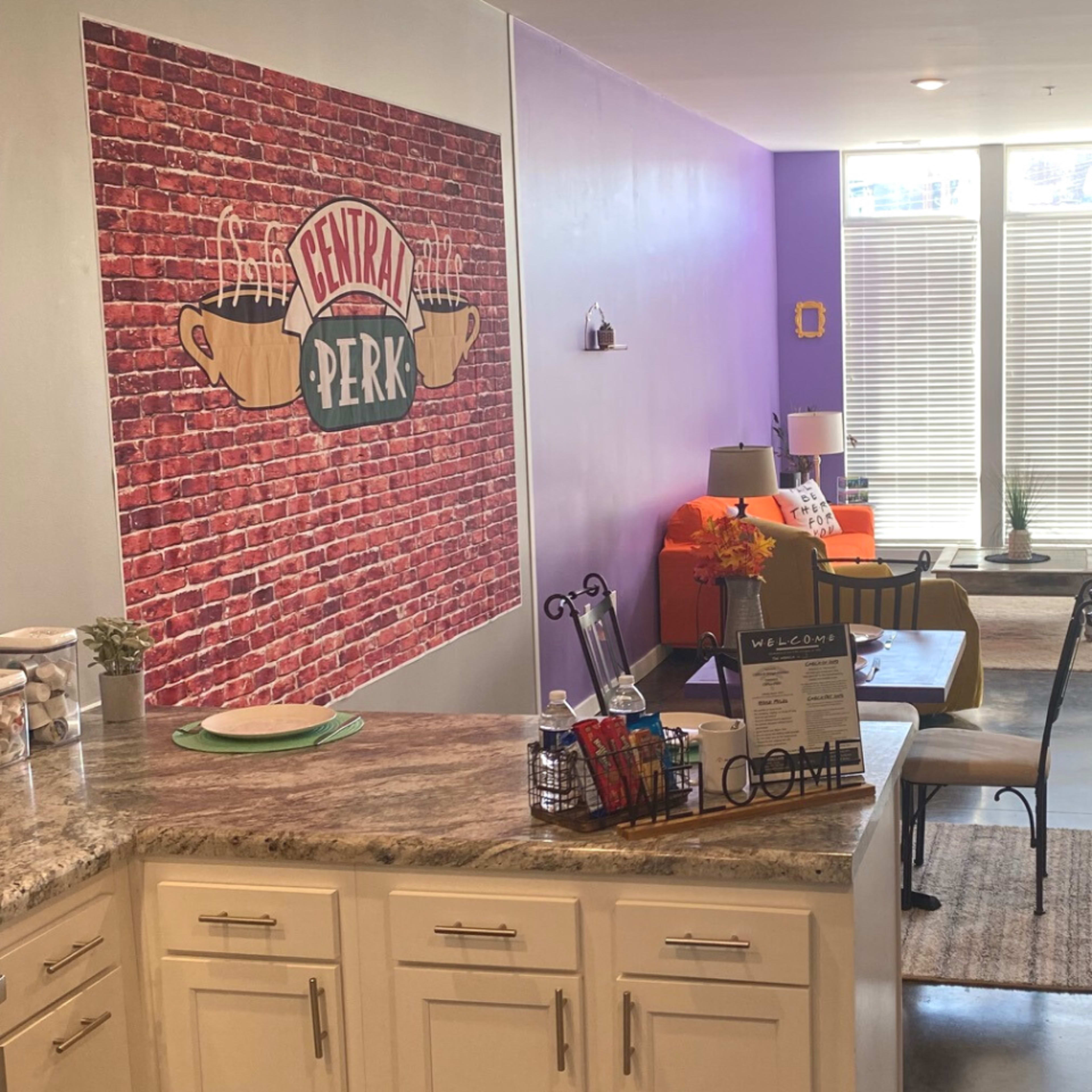 A modern living area with a kitchen and dining space featuring a brick accent wall and large windows allowing natural light.