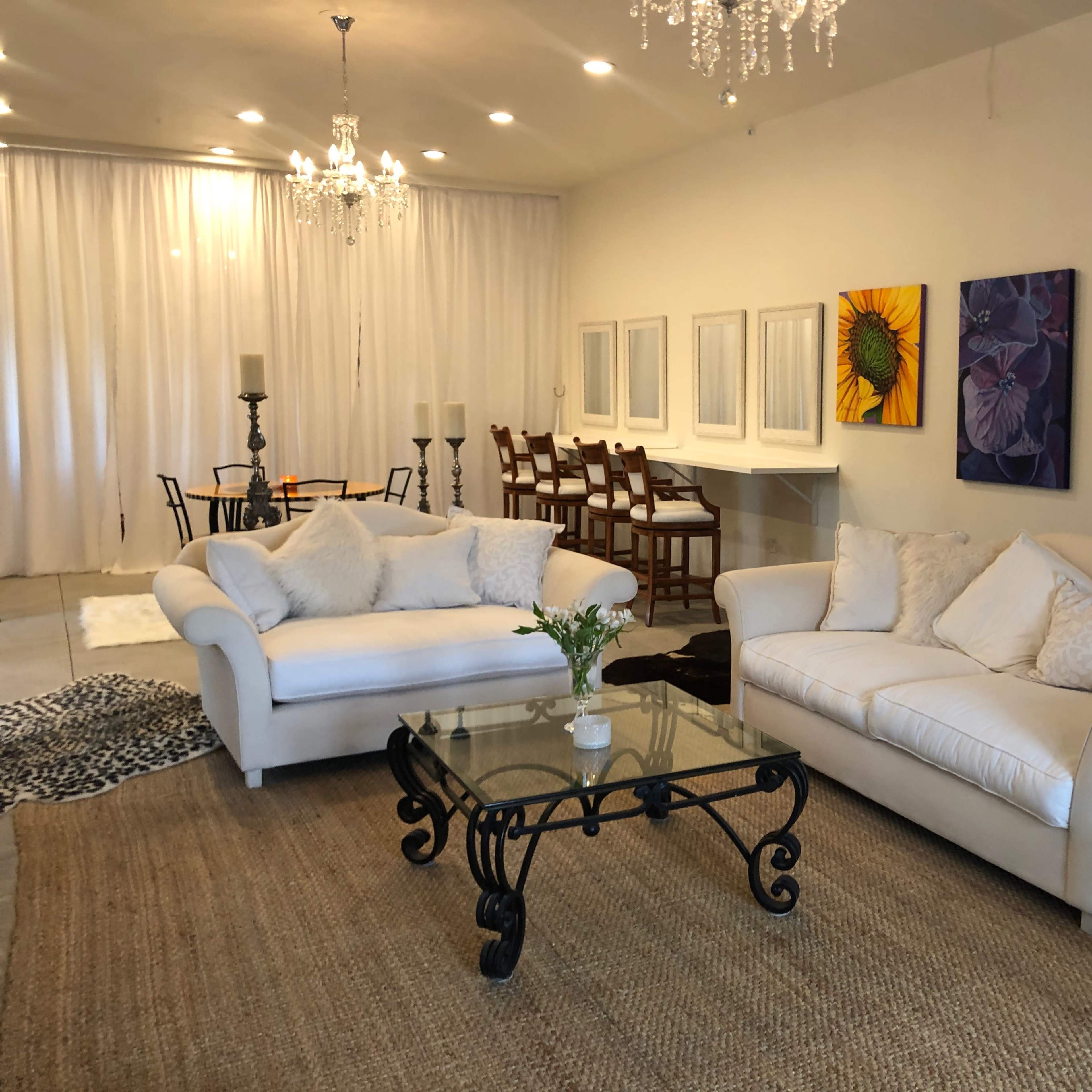 A modern living room with white sofas, a glass coffee table, and colorful artwork on the walls, leading into a dining area with a table and chairs.