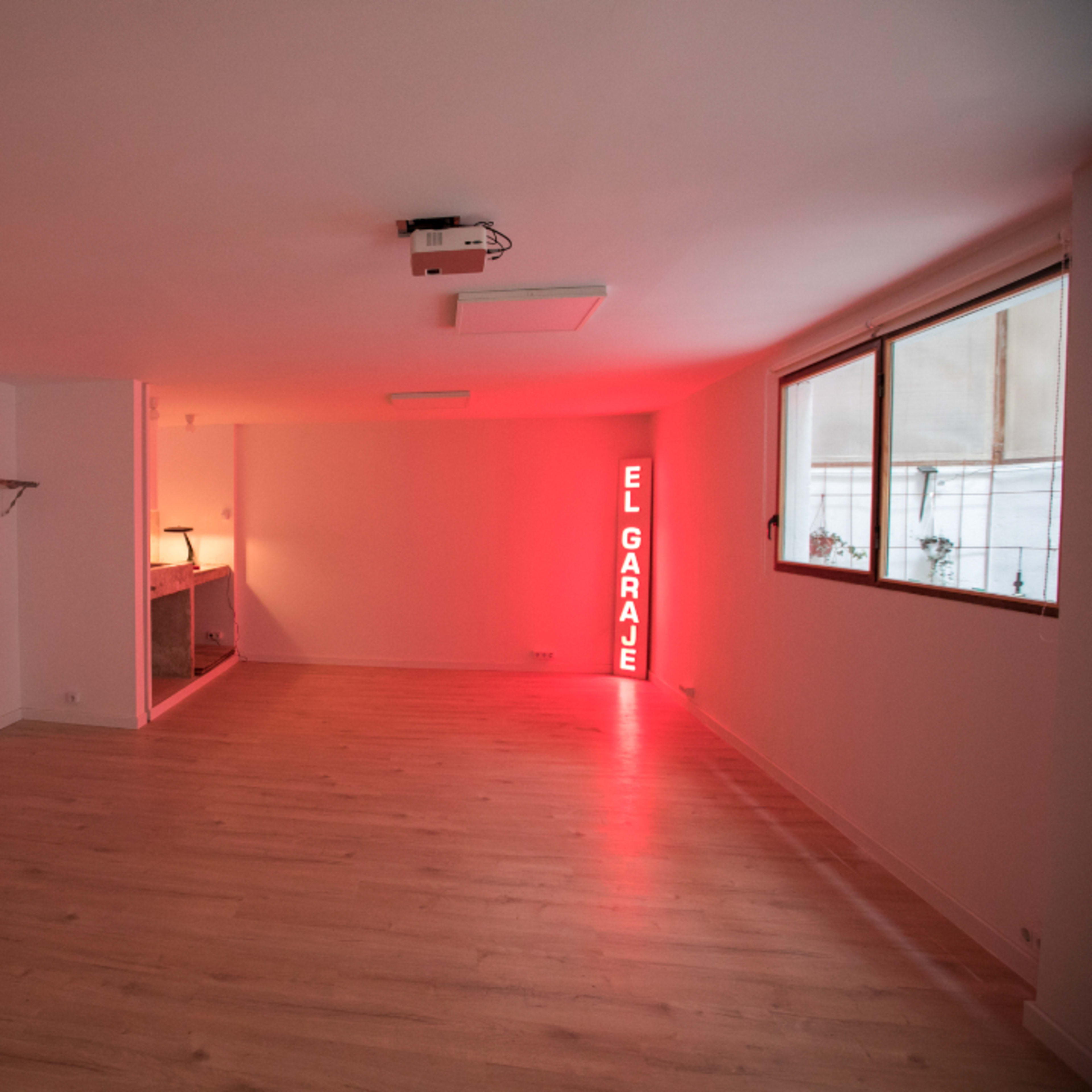 A spacious, empty room featuring wooden flooring, a guitar on the wall, a bar area, and a neon sign that says "EL GARAJE."