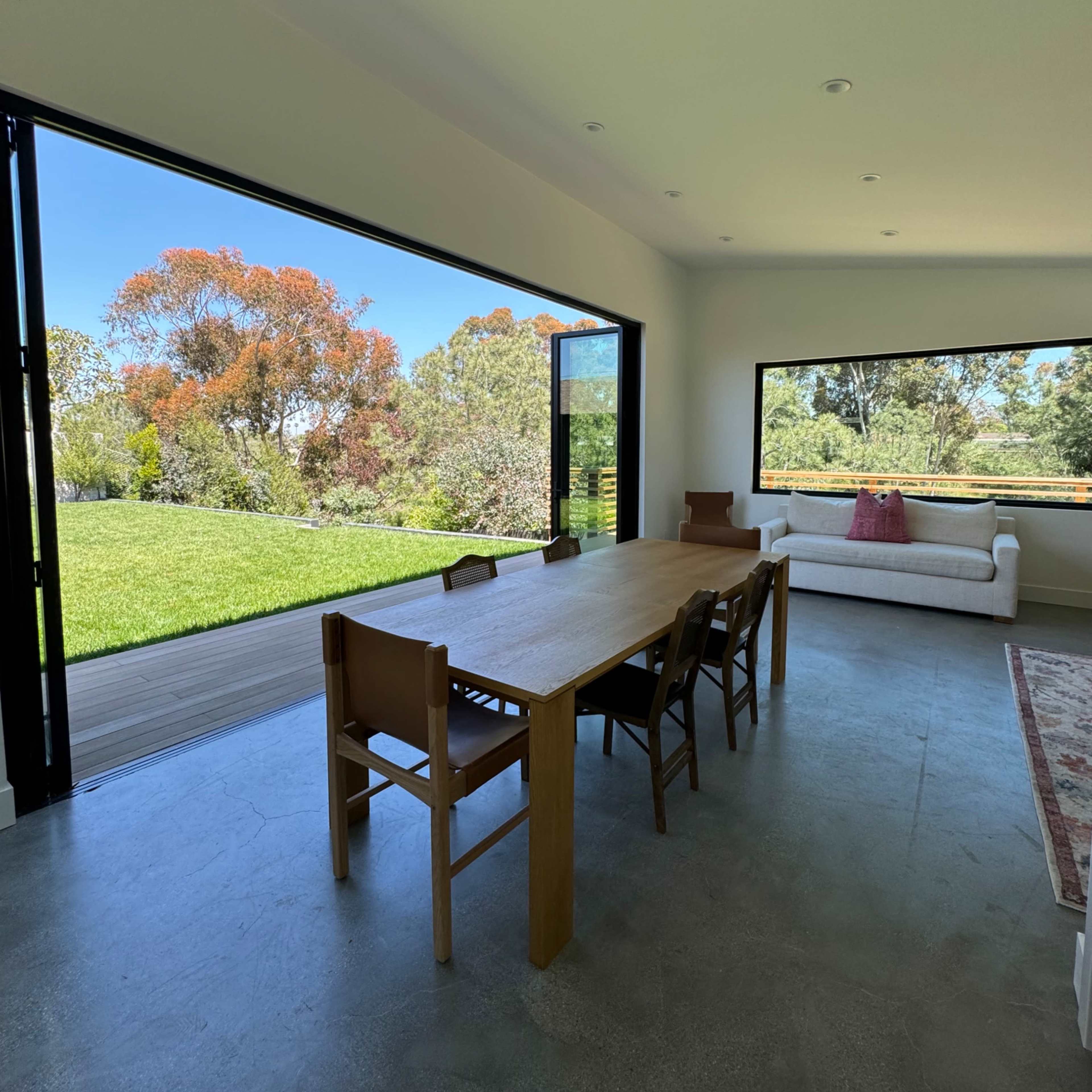 A spacious interior features a wooden dining table with chairs, leading to large sliding glass doors that open to a green landscape.