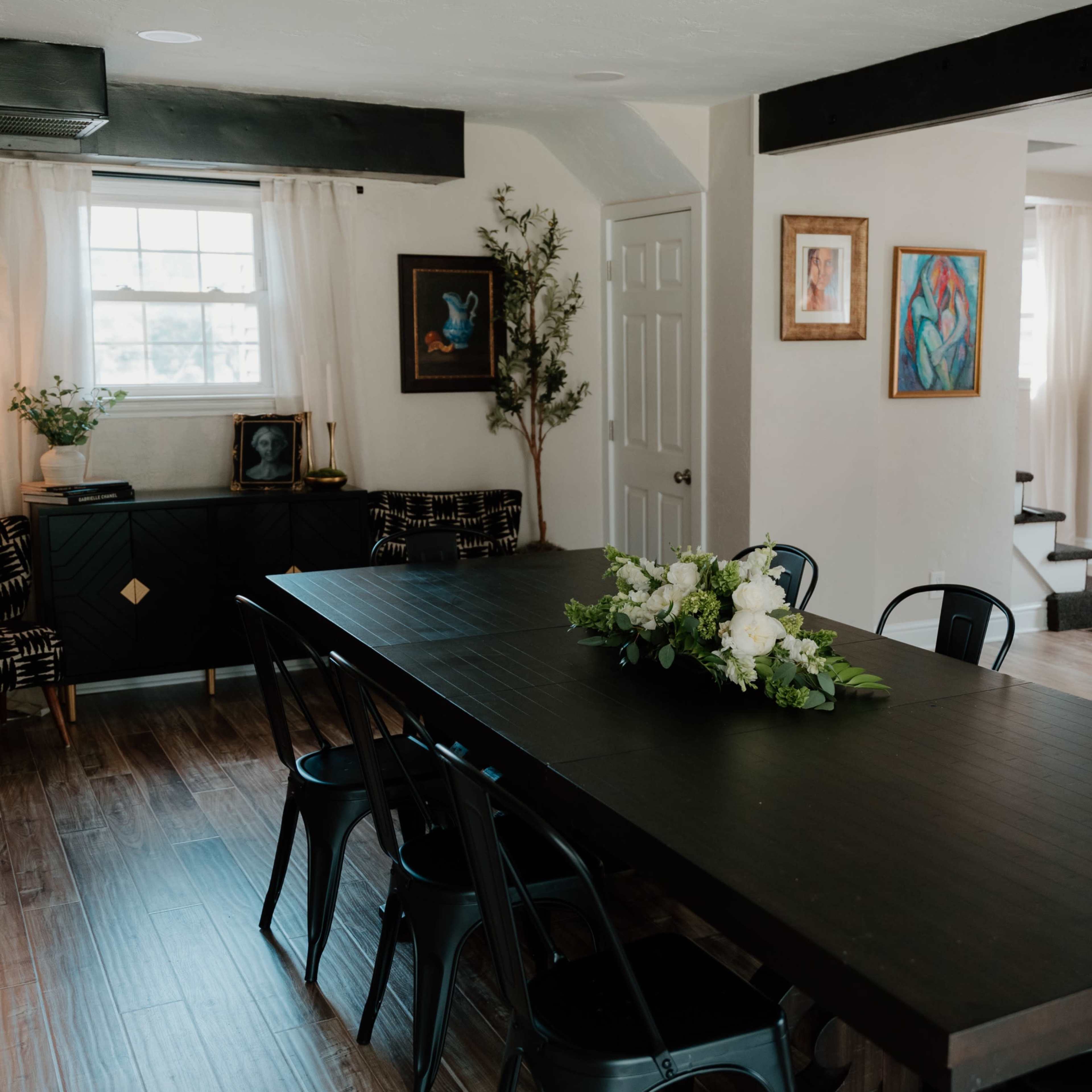 A long black dining table with metal chairs is set in a bright room featuring artwork and greenery.