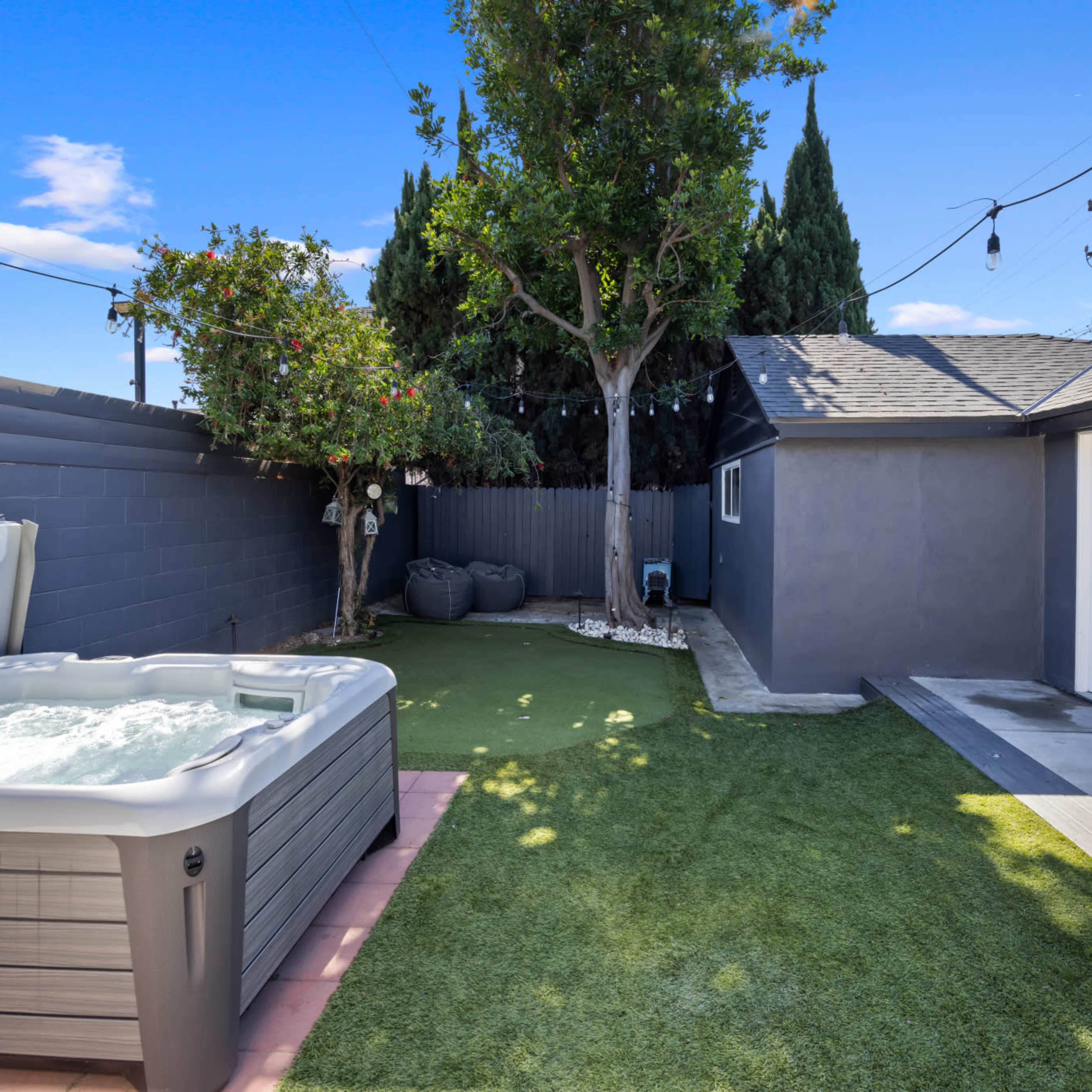 A backyard with a hot tub, artificial grass, a tree, and a small patio area alongside a gray house.