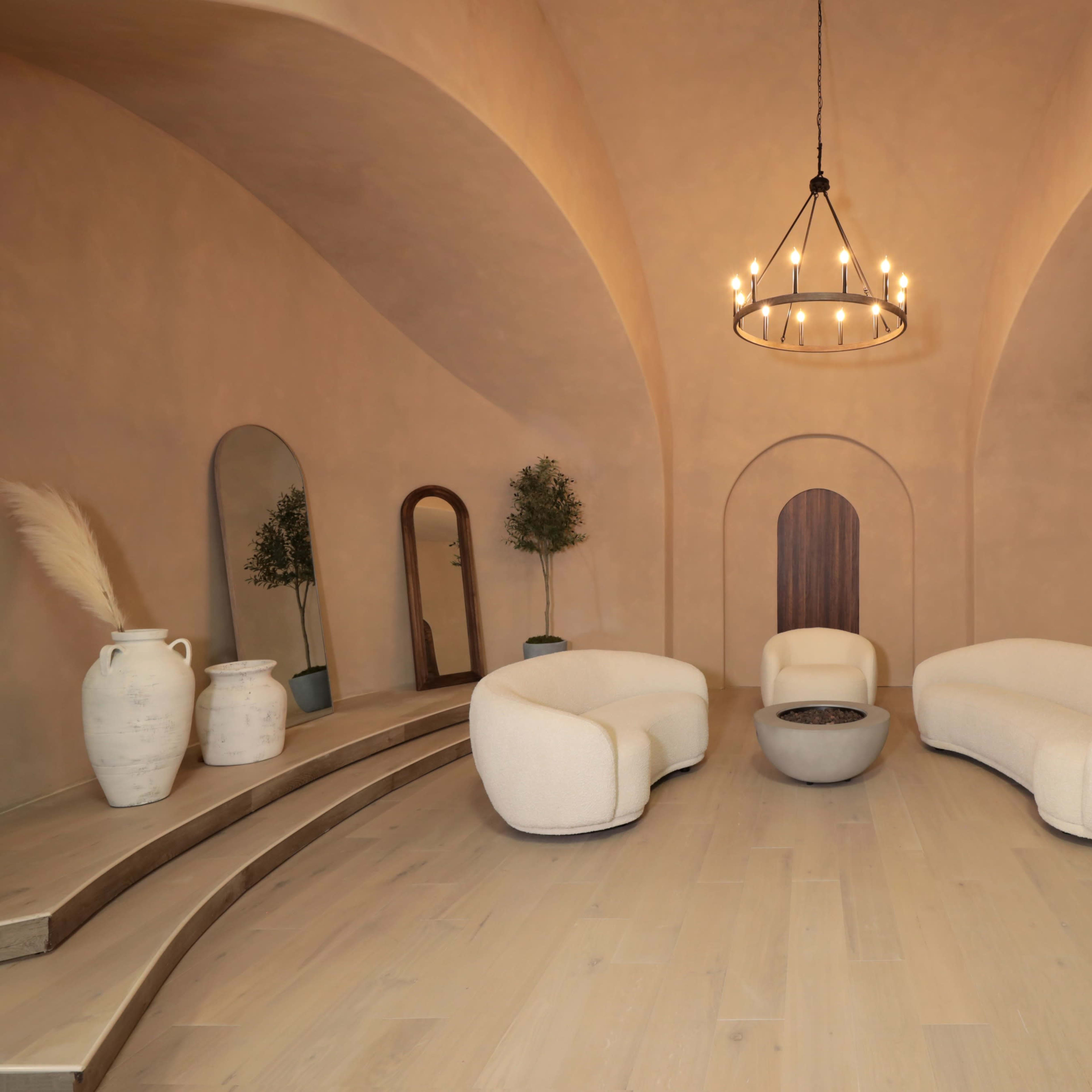 A spacious, minimalist interior with curved seating arrangements, a central table, and decorative elements such as mirrors and pottery set against a softly lit, arching wall.