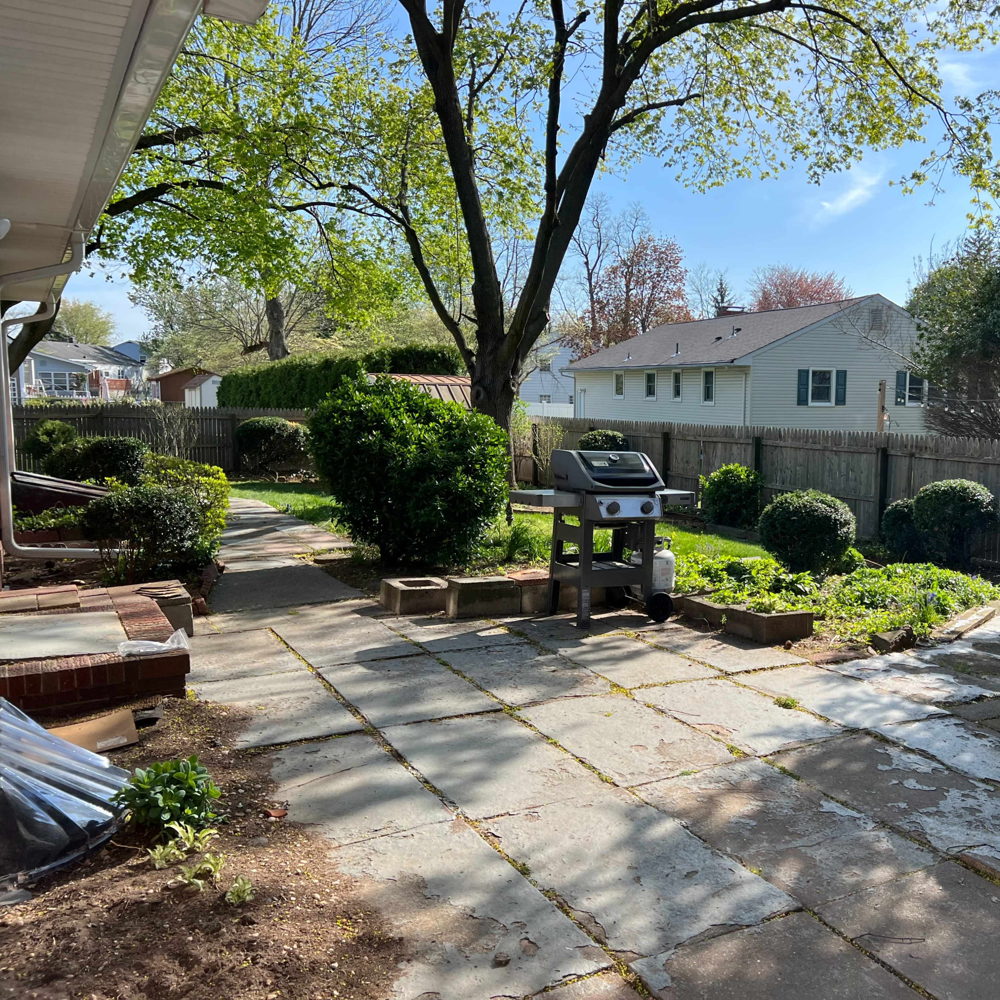 A patio area features a grill surrounded by greenery and well-maintained shrubbery, with houses visible in the background.