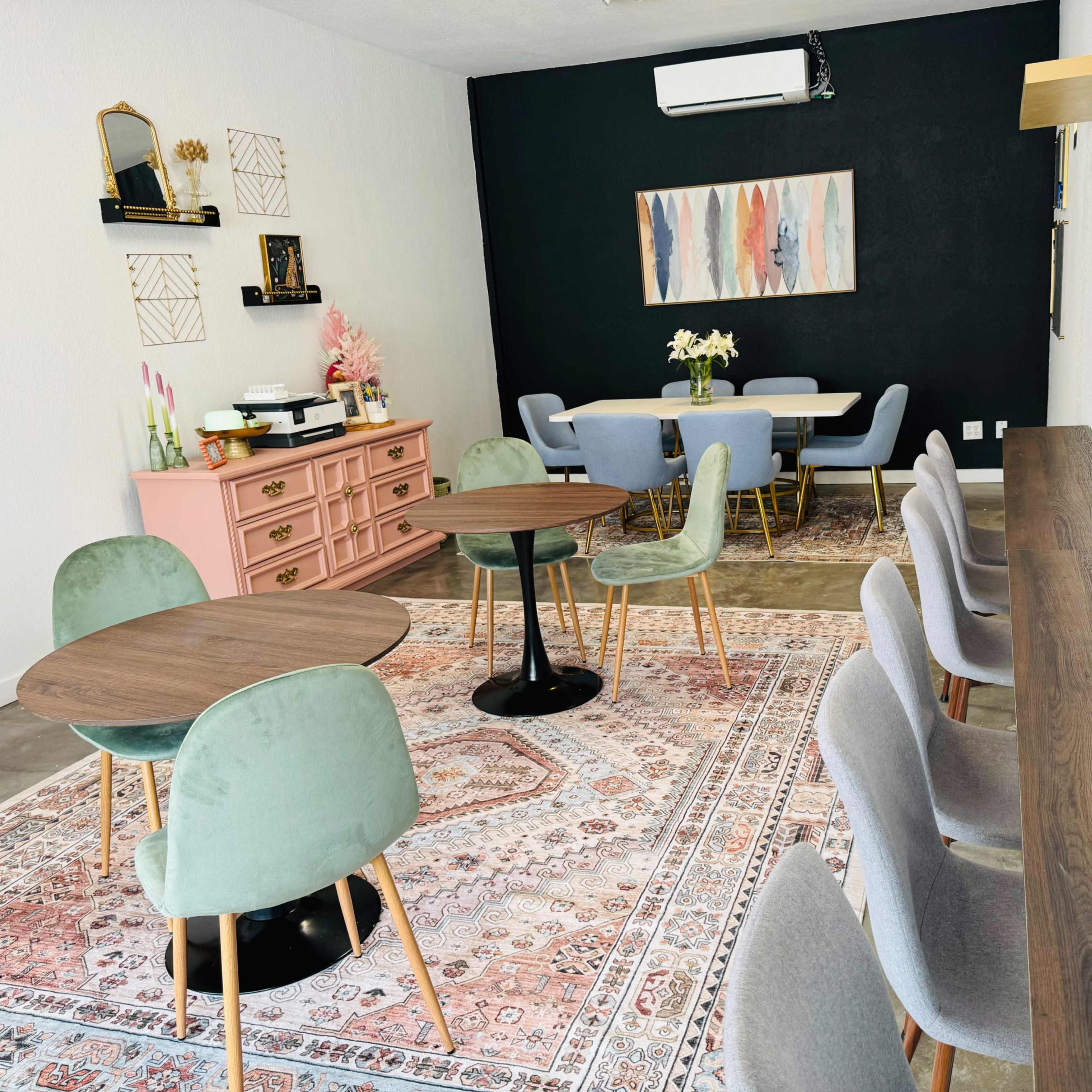 A modern interior space featuring a set of tables and chairs arranged around a decorative rug, with a pink dresser and a wall-mounted artwork in the background.