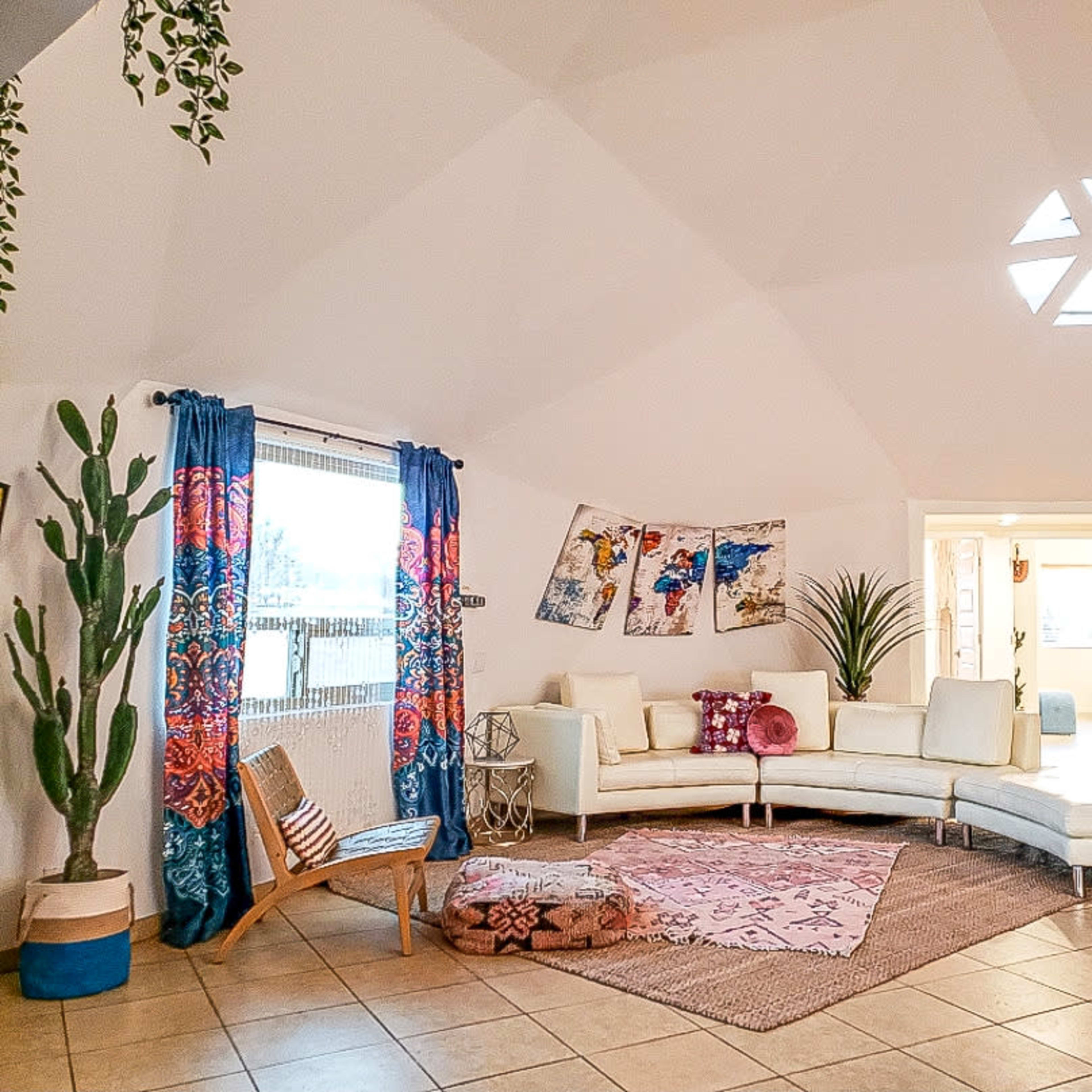 A stylish living room with a curved white sofa, decorative plants, colorful curtains, and artwork on the walls.