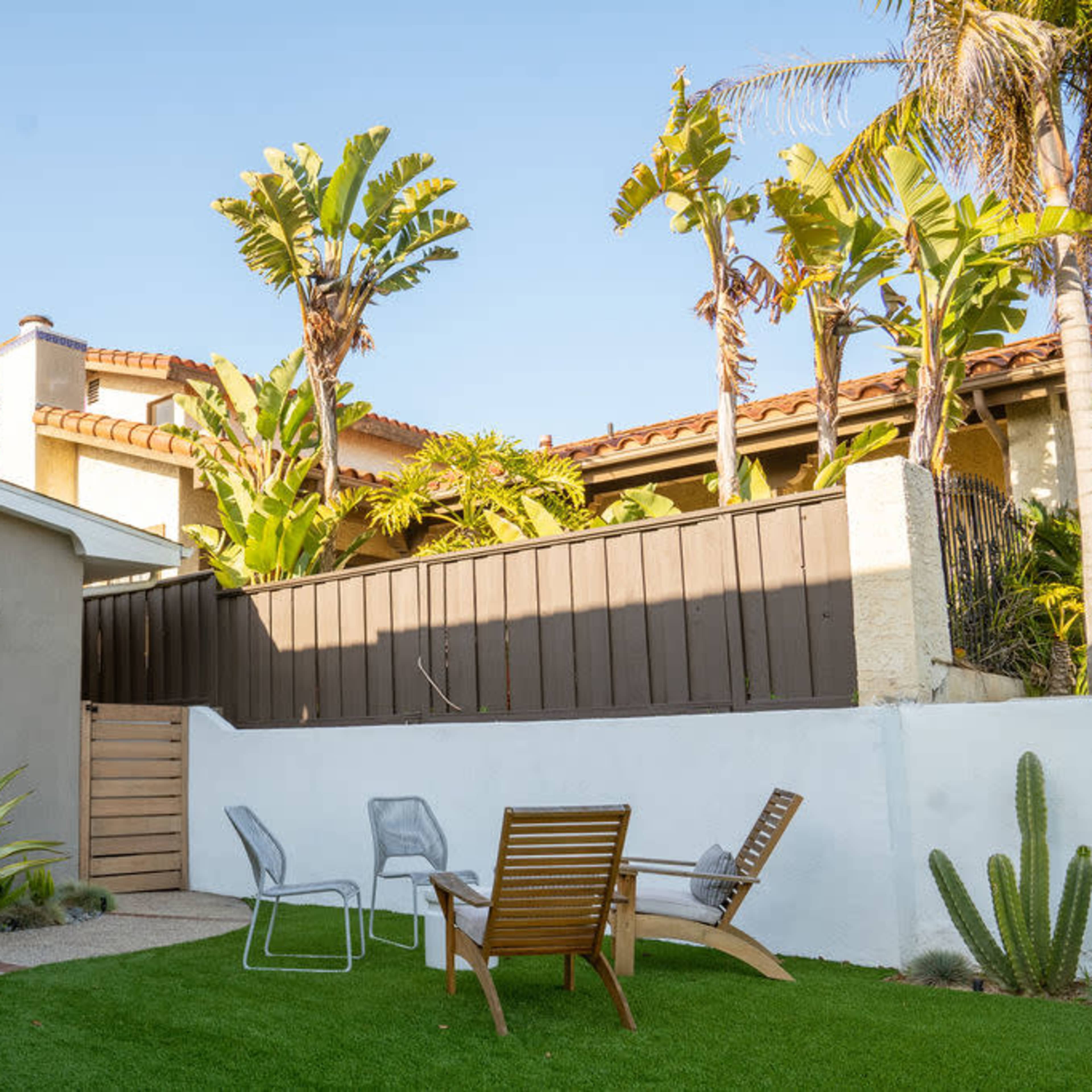 A modern backyard features a green lawn, a seating area with two chairs and a side table, surrounded by tall plants and a wooden fence.