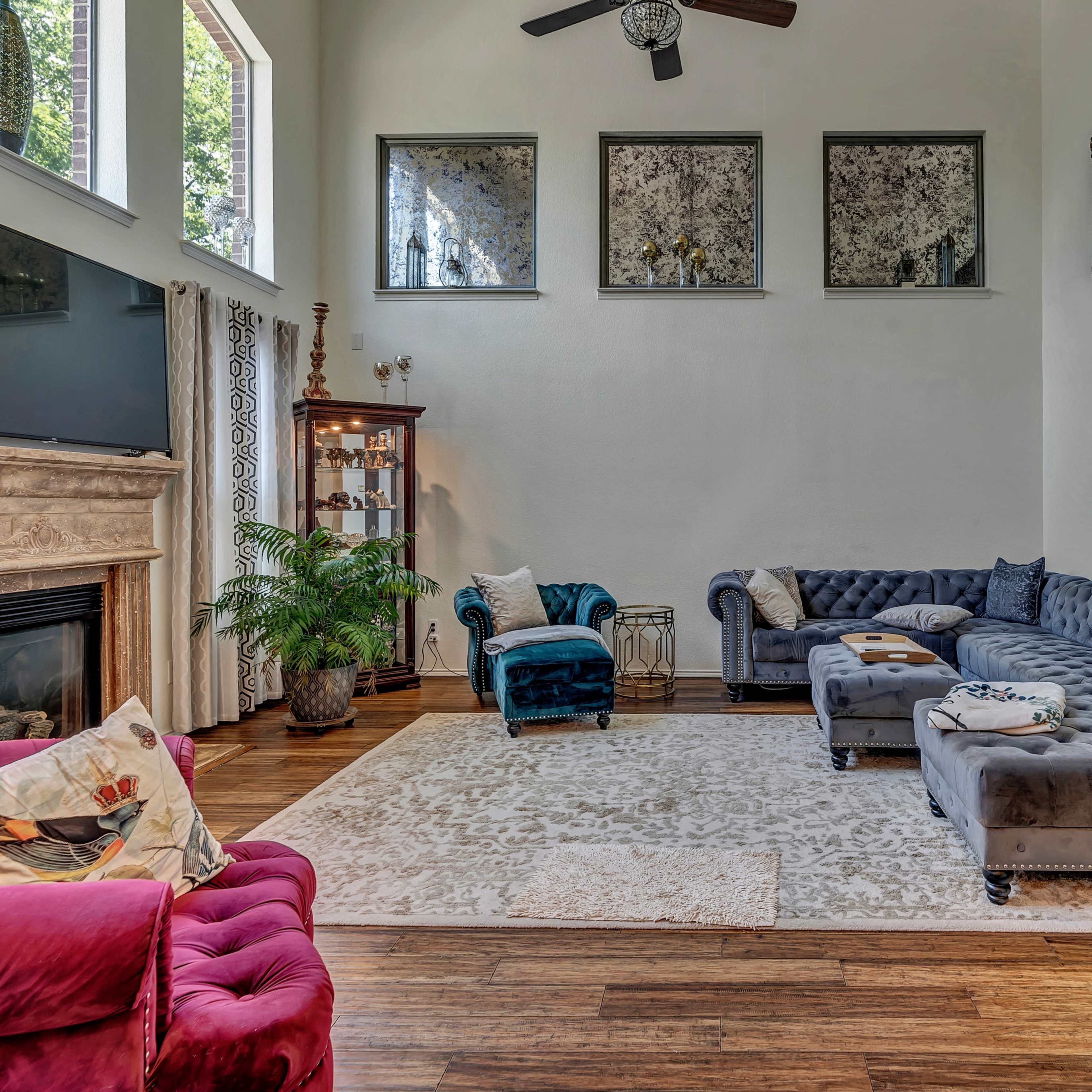 The living room features a sectional sofa, a plush armchair, and a decorative rug on wooden flooring, with large windows allowing natural light to enter.