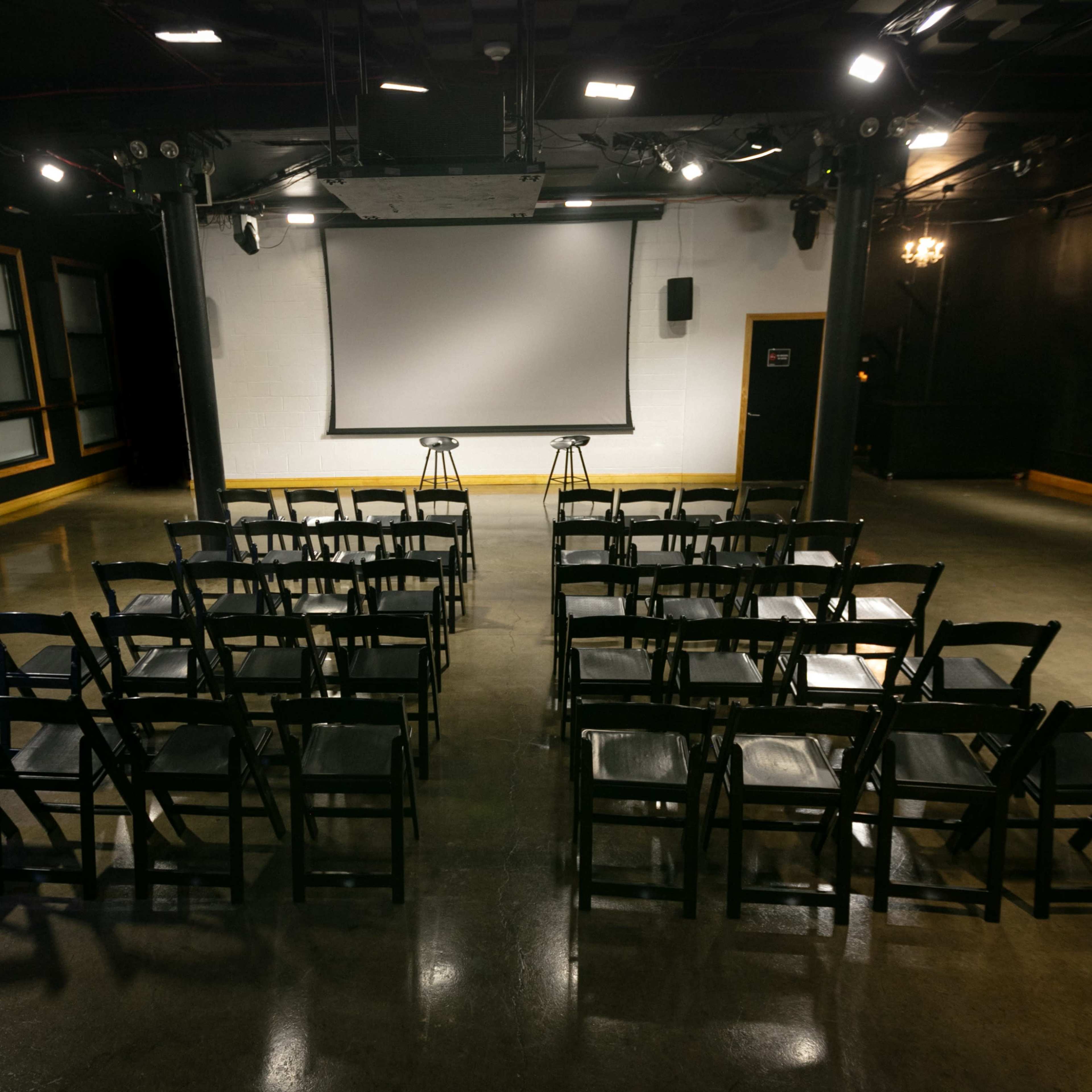 A presentation space is set up with rows of black chairs facing a screen and two stools.