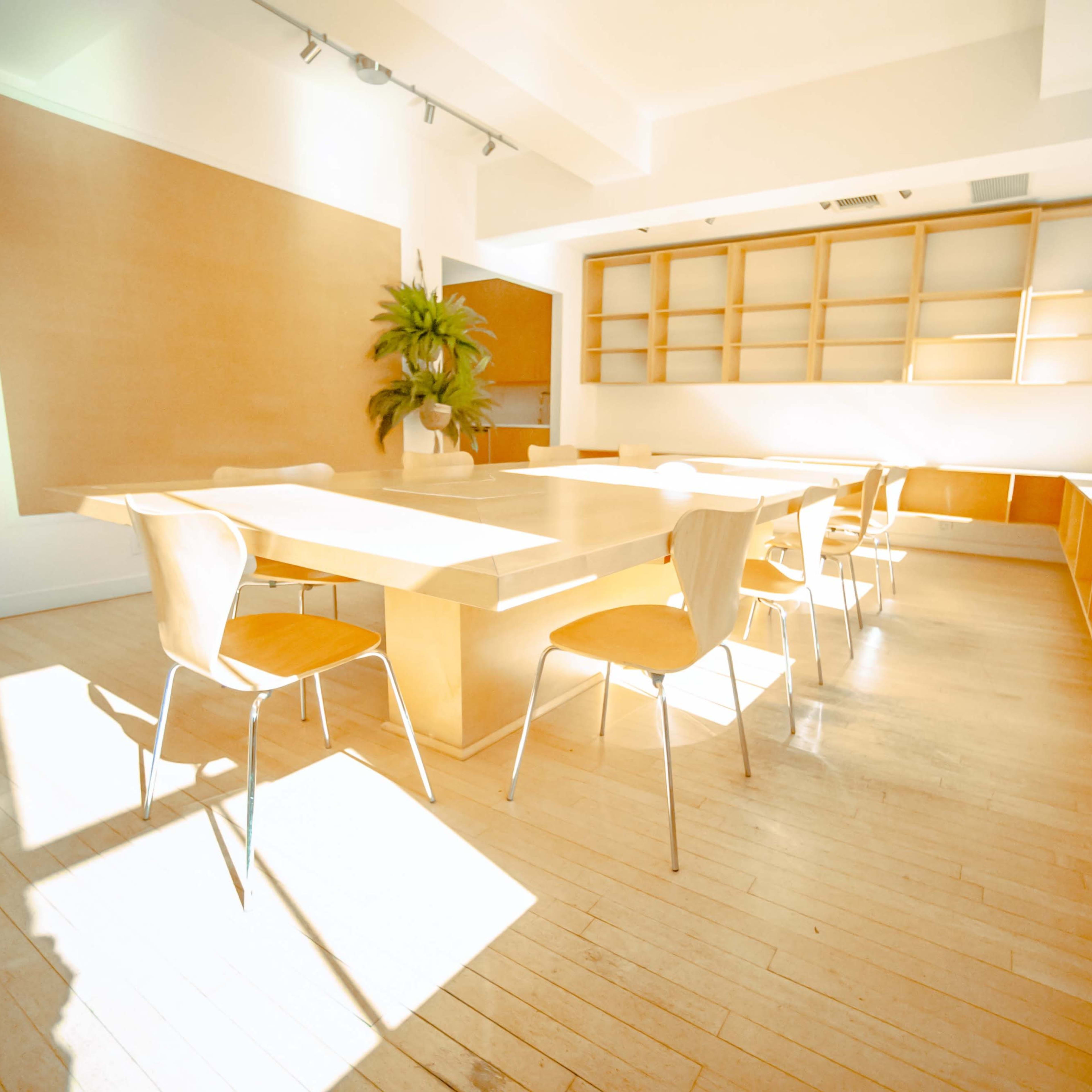 A bright, minimalist room features a large wooden table surrounded by several chairs and a plant, with light streaming through large windows.