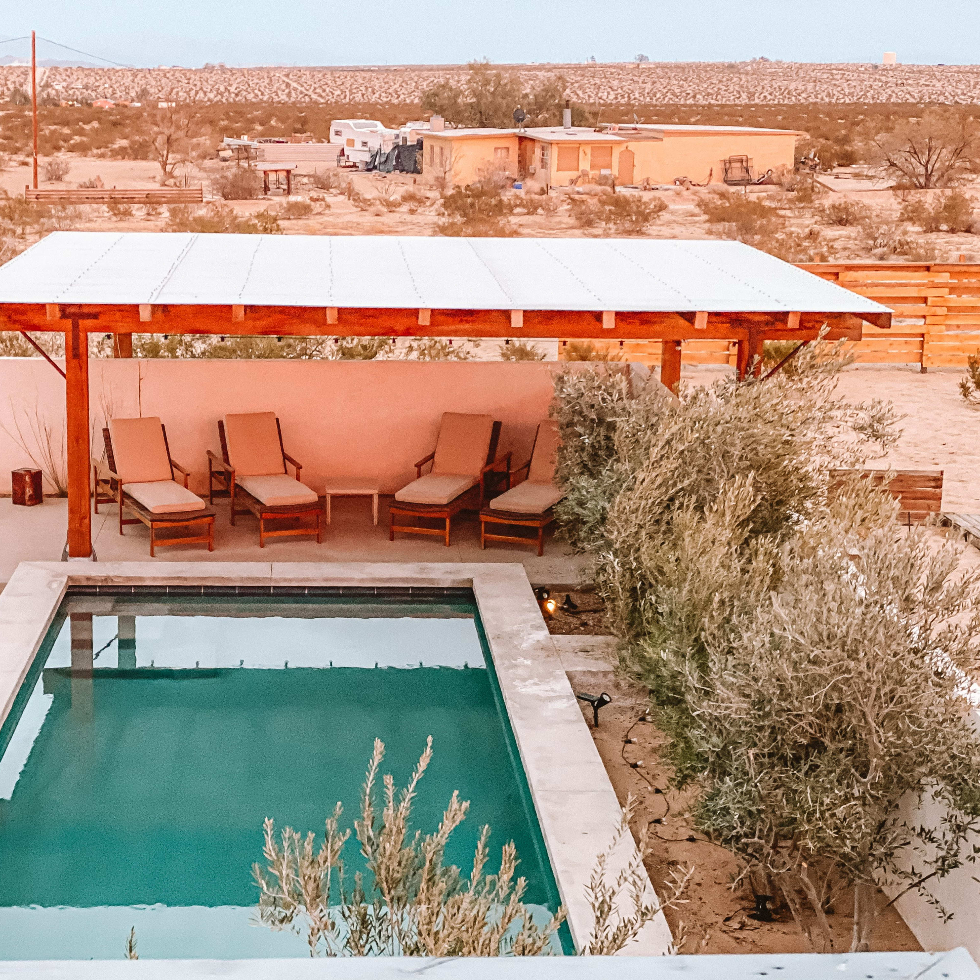 A pool area with lounge chairs under a shaded structure, surrounded by desert vegetation and distant buildings.